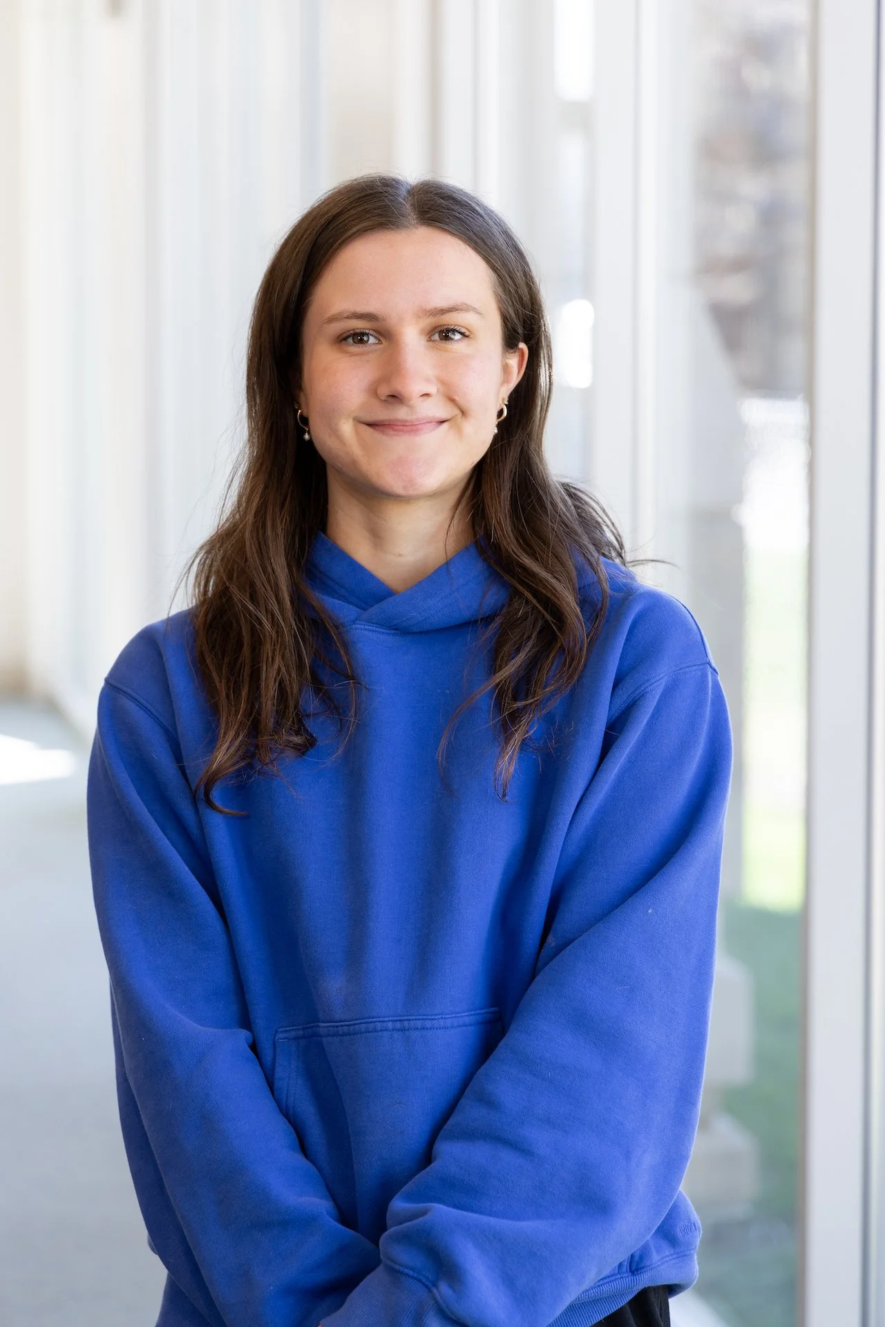 A young woman wearing a blue hoodie, standing indoors near large windows, smiling at the camera.