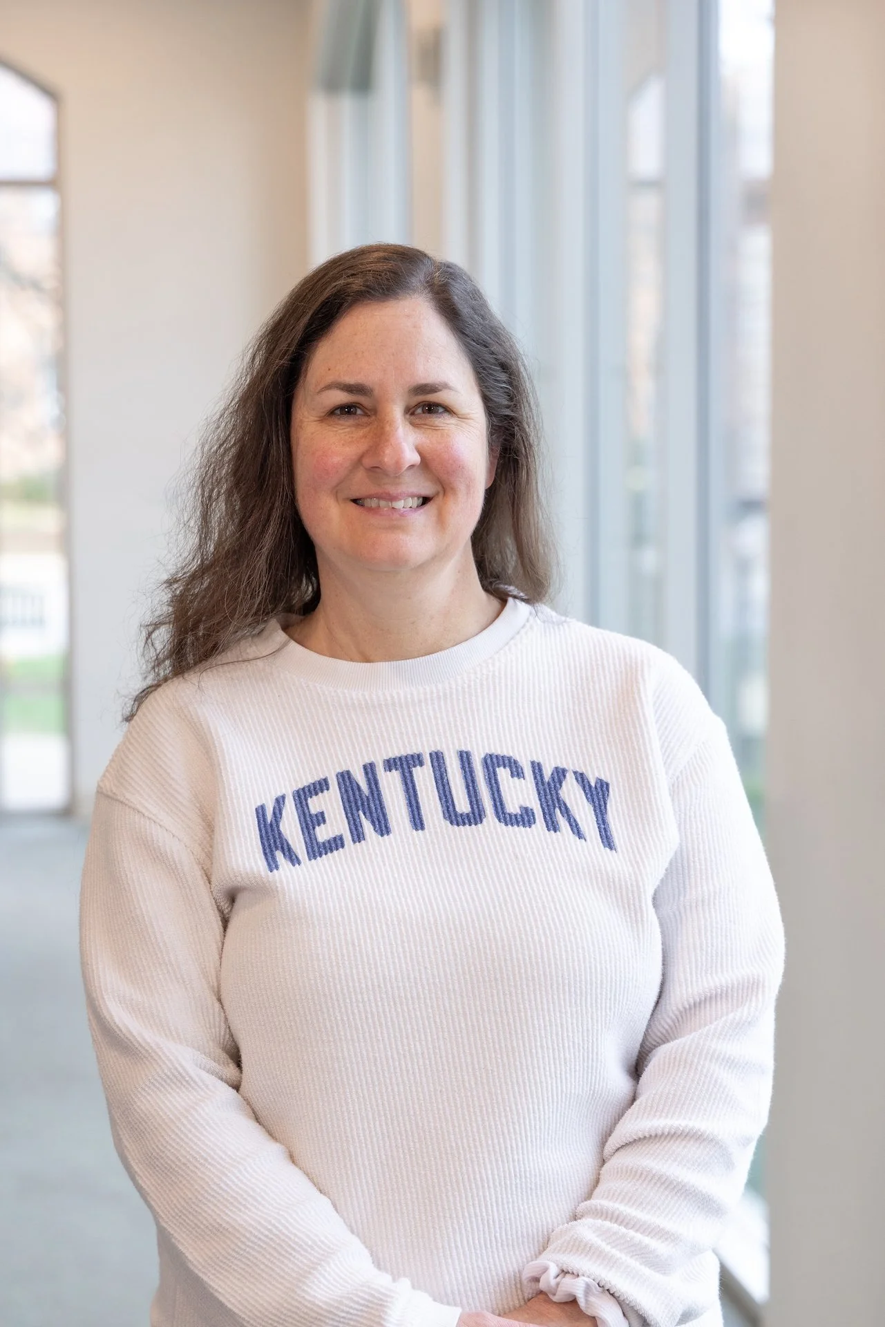 Woman with brown hair smiling, wearing a cream sweater with 'KENTUCKY' written in blue letters, standing indoors near windows.