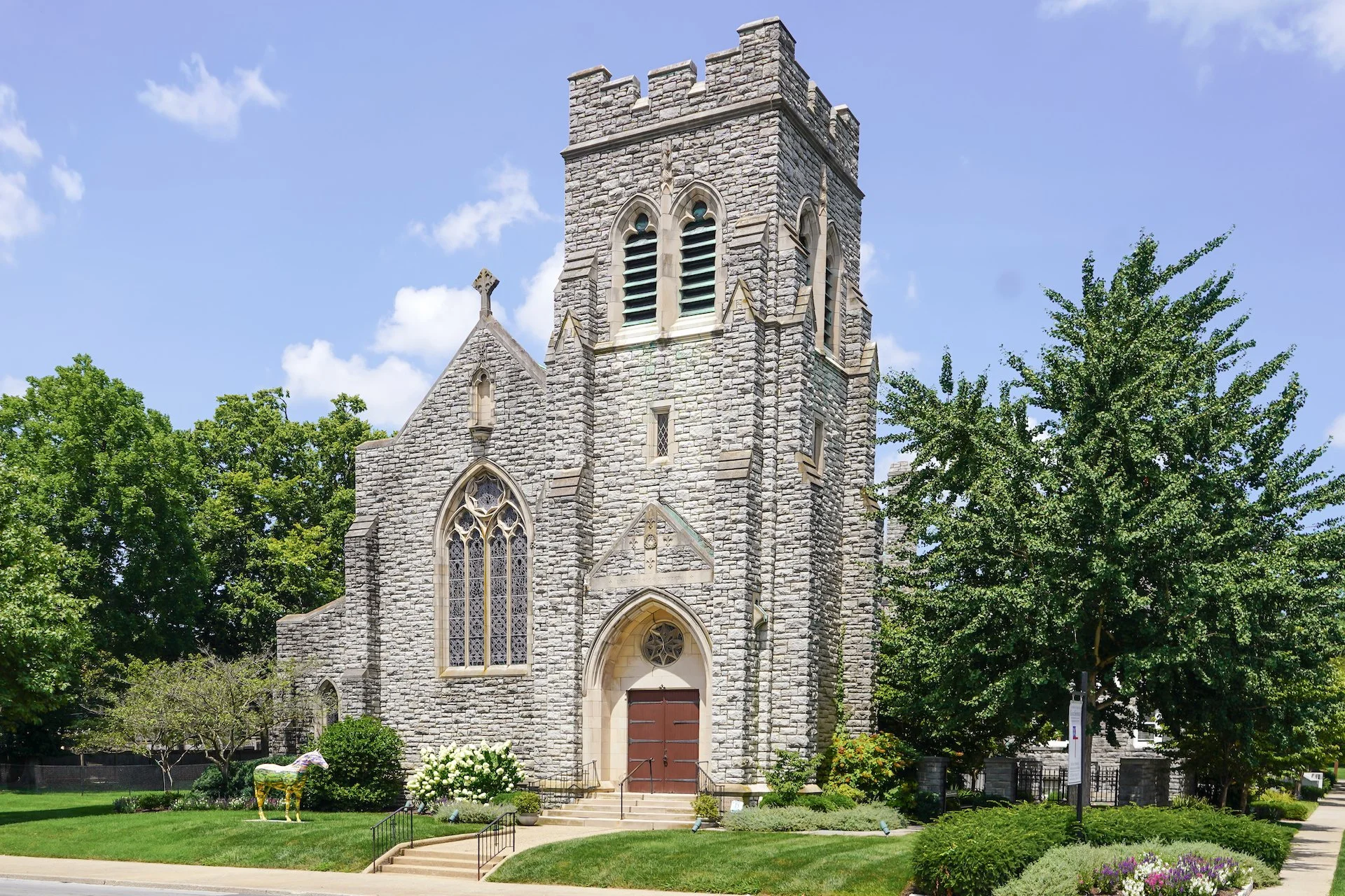 A stone church with tall, arched stained glass windows and a bell tower, surrounded by green trees and a manicured lawn under a blue sky with some clouds.
