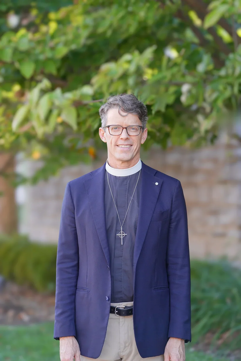 A man wearing glasses, a black clergy shirt with a white clerical collar, a navy blazer, and a large cross necklace, standing outdoors in front of green foliage and a brick wall.