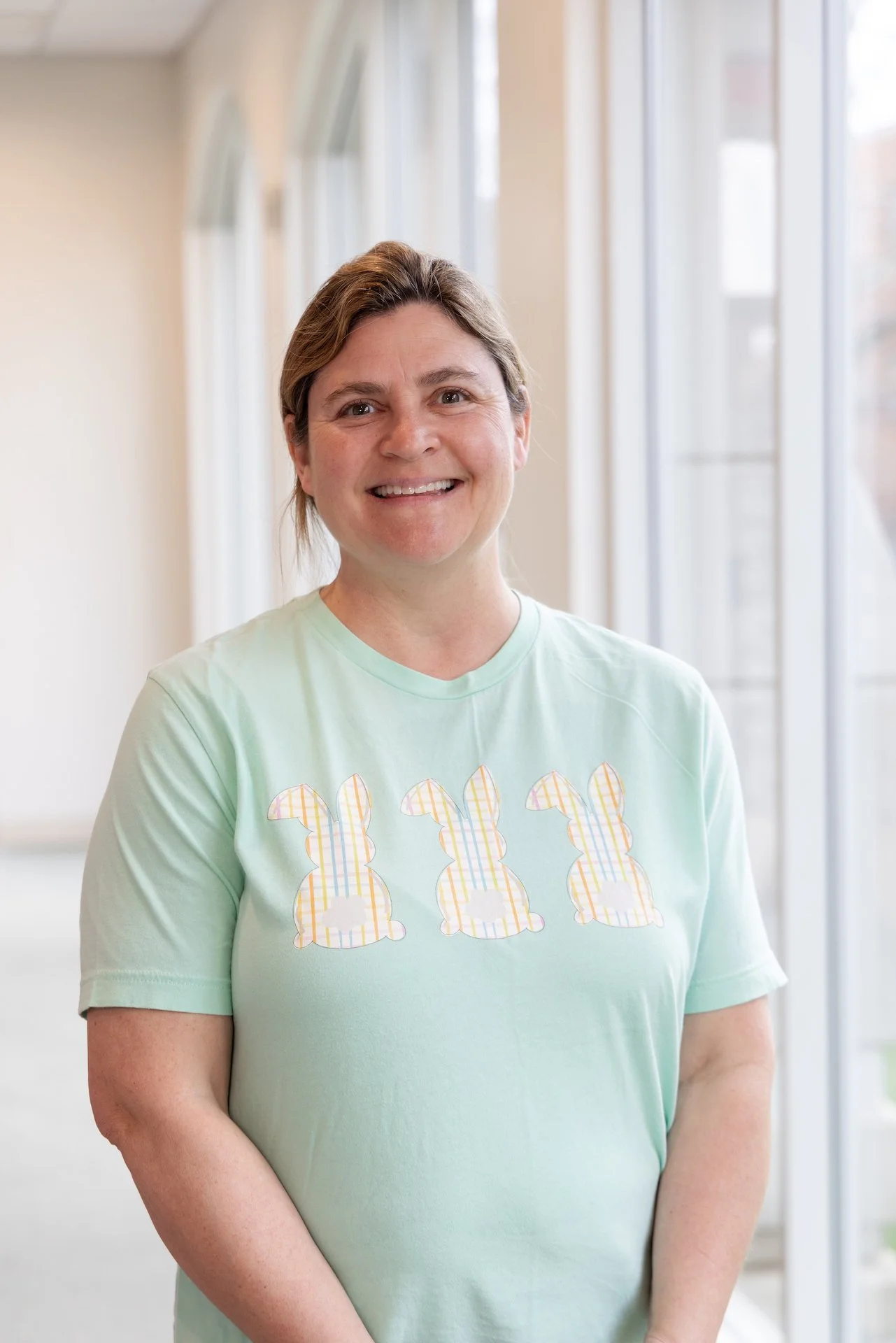 A smiling woman wearing a light green t-shirt with three bunny illustrations, standing in front of a row of windows.