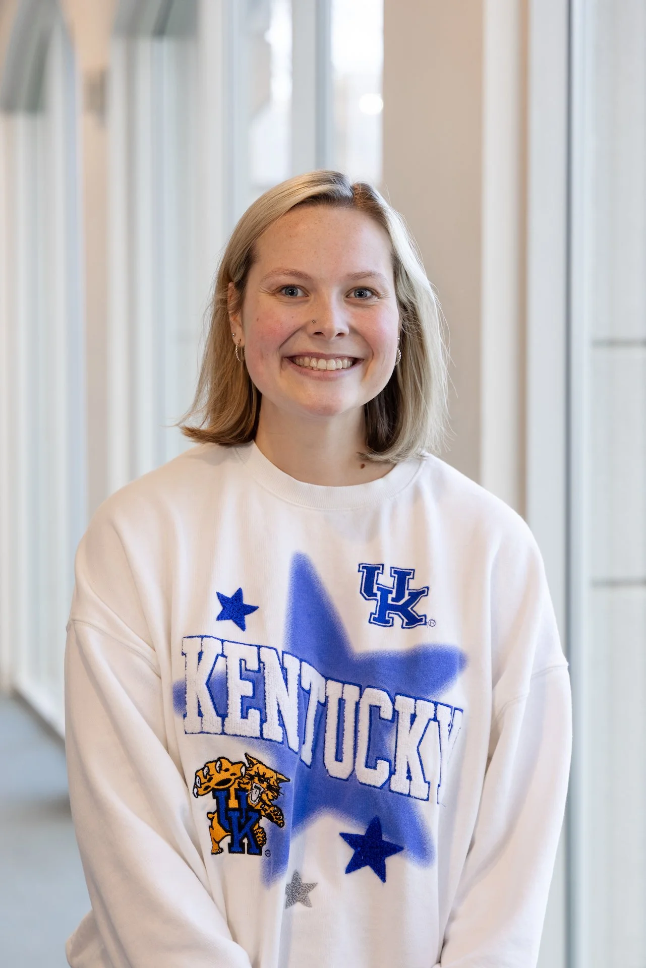 A young woman with shoulder-length blonde hair smiling, wearing a white sweat shirt with Kentucky Wildcats logos and text, standing in front of windows.