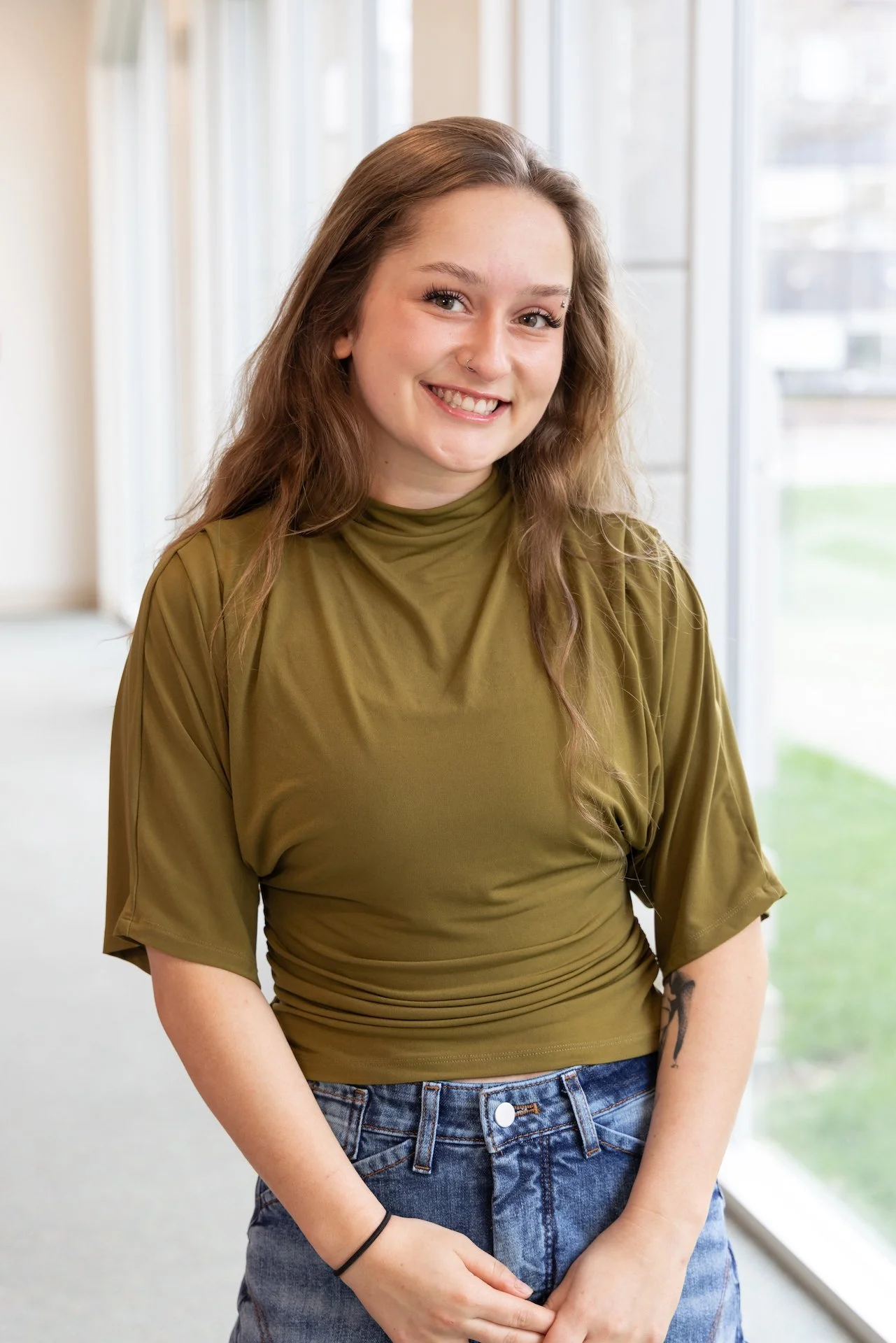 A young woman with long brown curly hair, smiling, wearing an olive green top and blue jeans, standing inside near large windows.