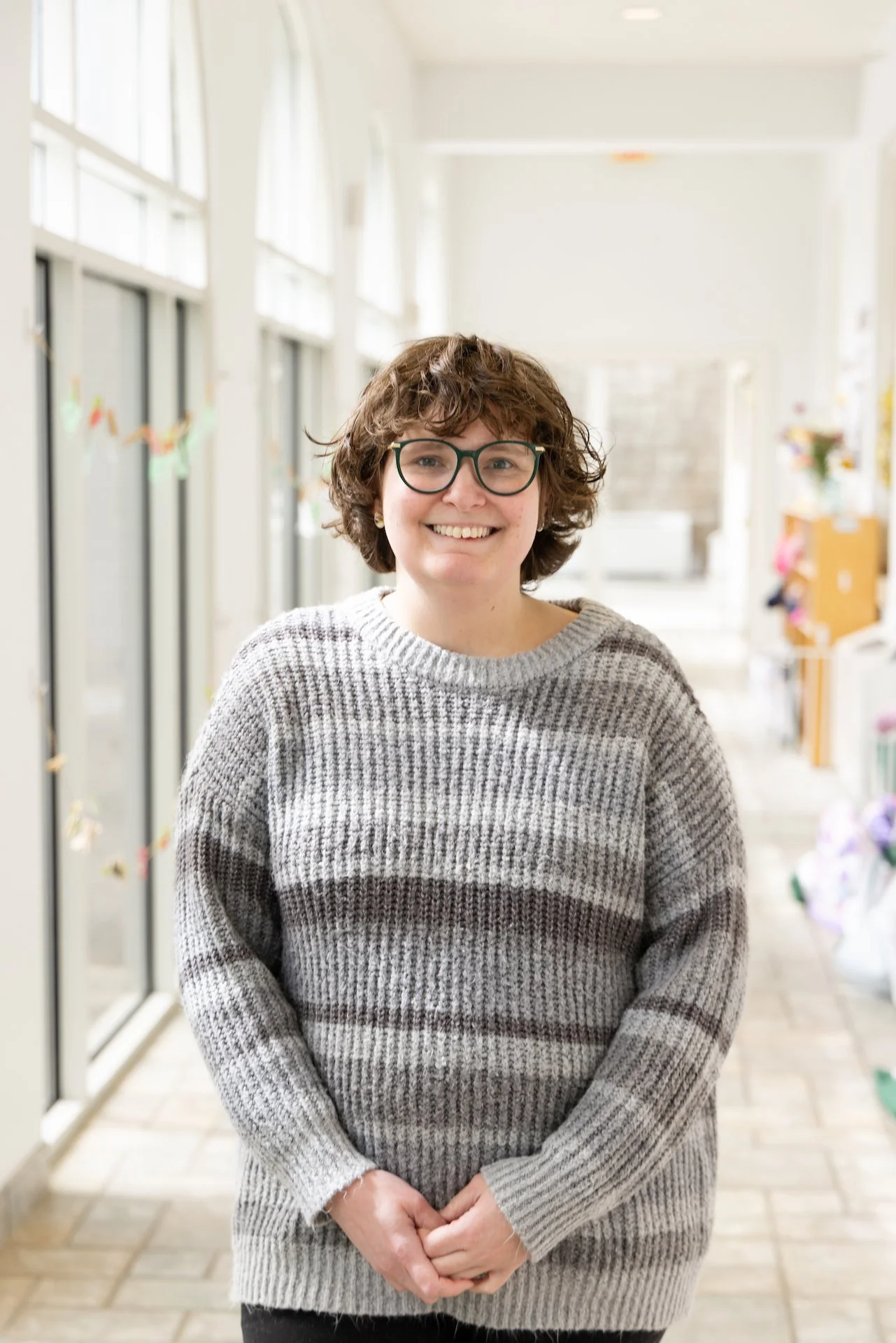 A woman with short curly hair and glasses, smiling and wearing a gray striped sweater, standing in a brightly lit hallway.