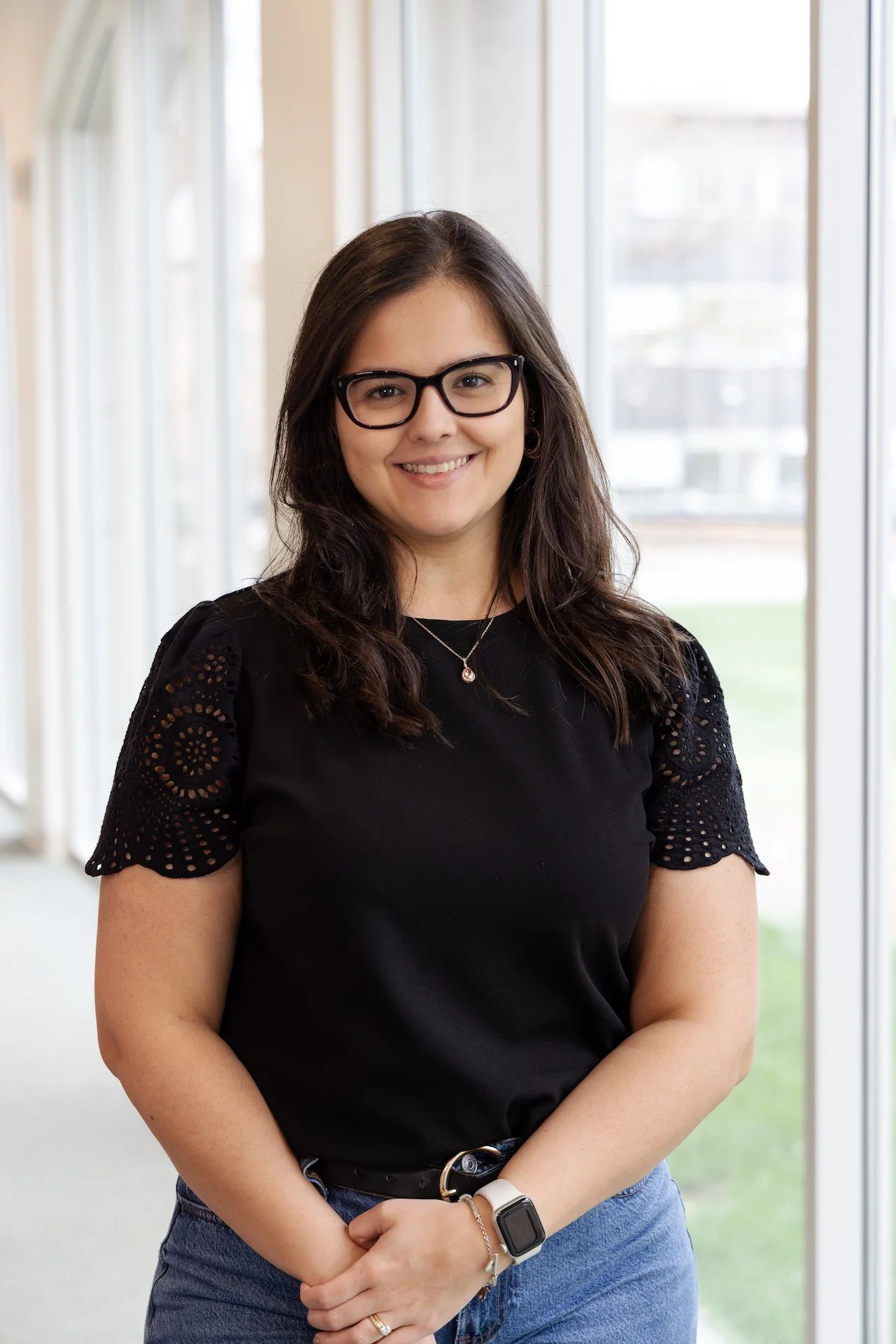 A woman with dark hair, wearing glasses, a black top with eyelet details on the sleeves, blue jeans, a white smartwatch, and a necklace, standing indoors near large windows with a view of a building and greenery outside.