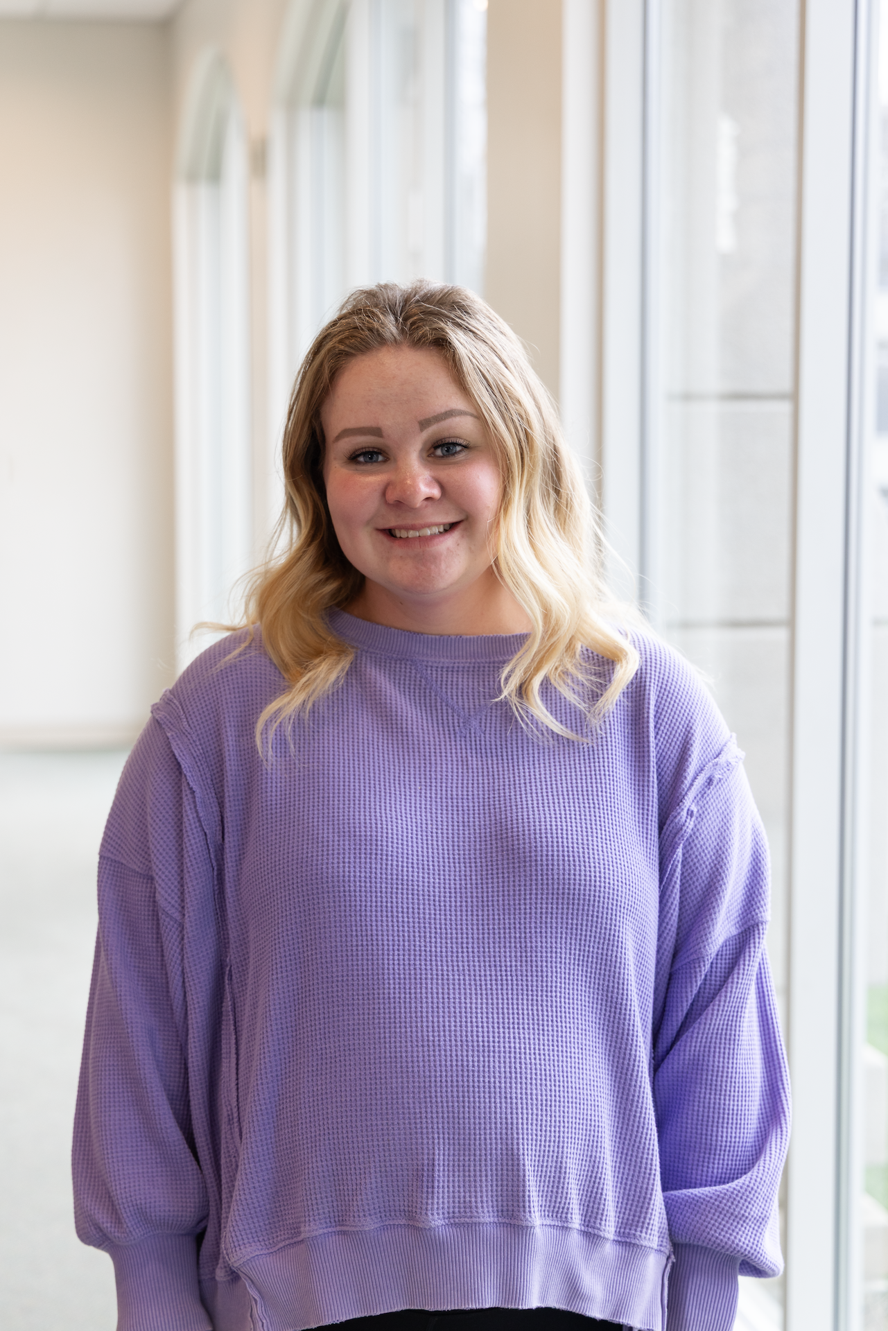 A young woman with blonde hair wearing a purple sweater, standing indoors near large windows, smiling at the camera.