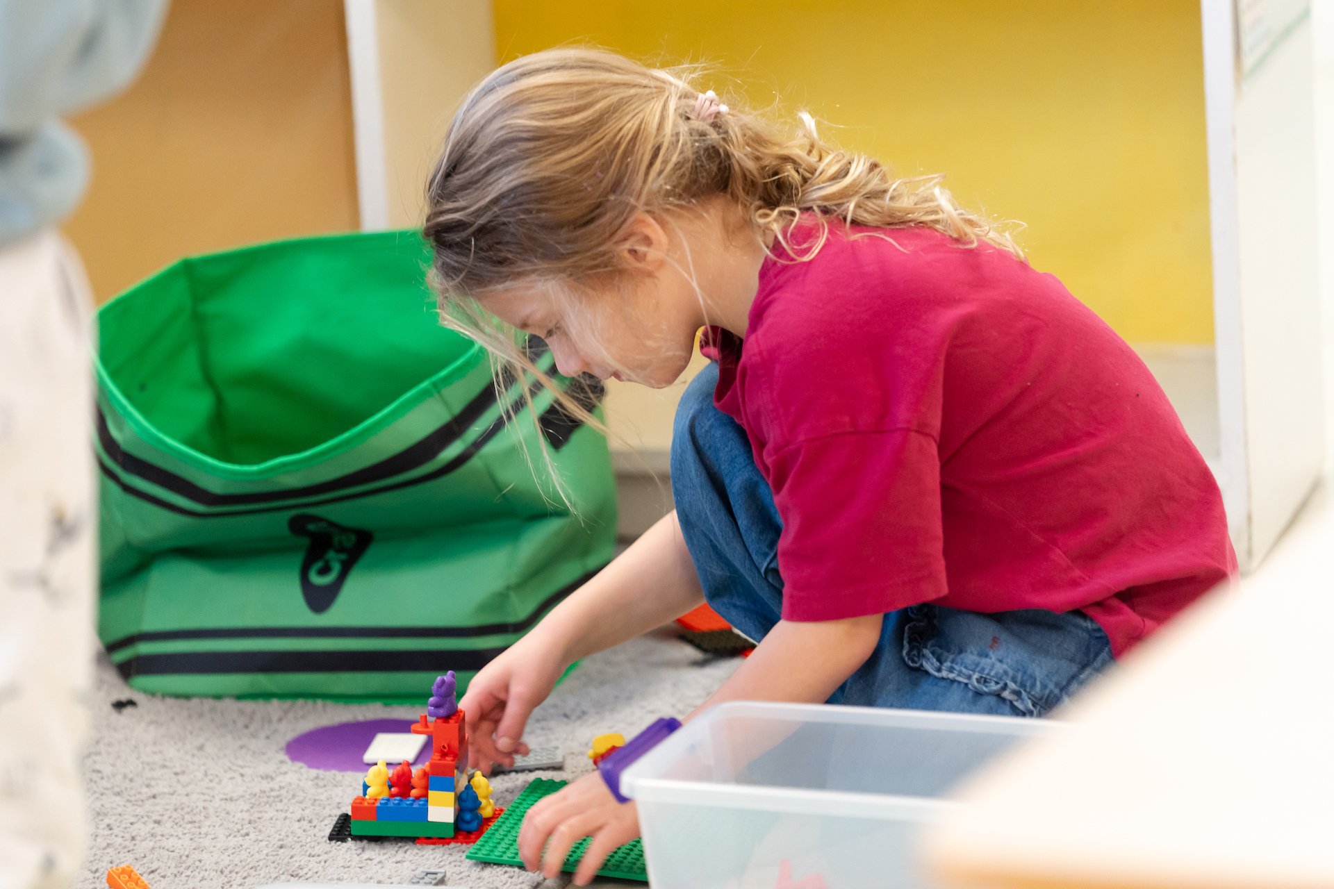 A young girl with blond hair in a ponytail, wearing a red T-shirt and blue jeans, is sitting on the carpet playing with colorful LEGO bricks. She is focused on building something, with a large green and black striped bag nearby.