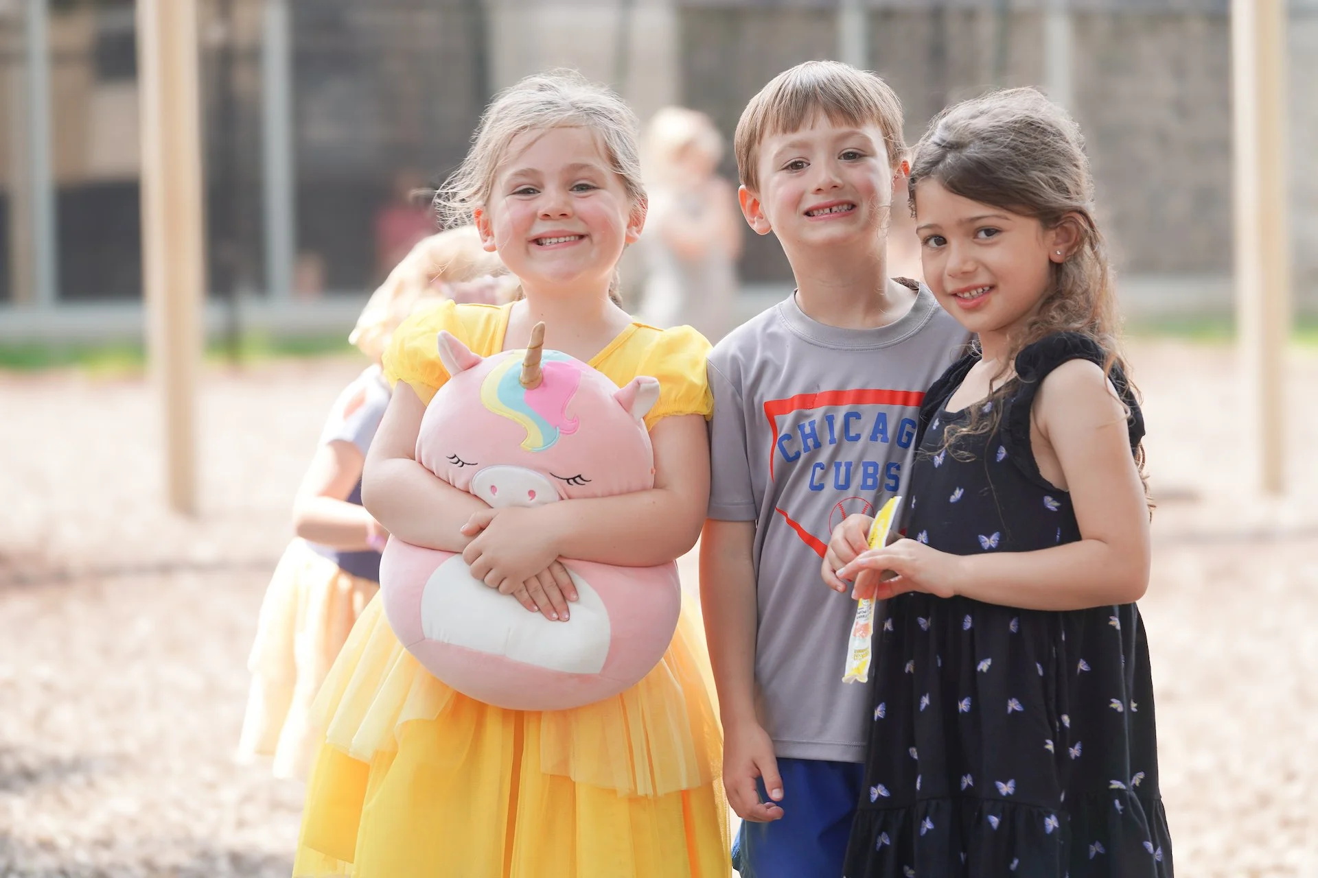 Smiling children standing outdoors, one girl in a yellow dress holding a unicorn plush toy, a boy wearing a Chicago Cubs shirt, and another girl in a black dress with a butterfly pattern.