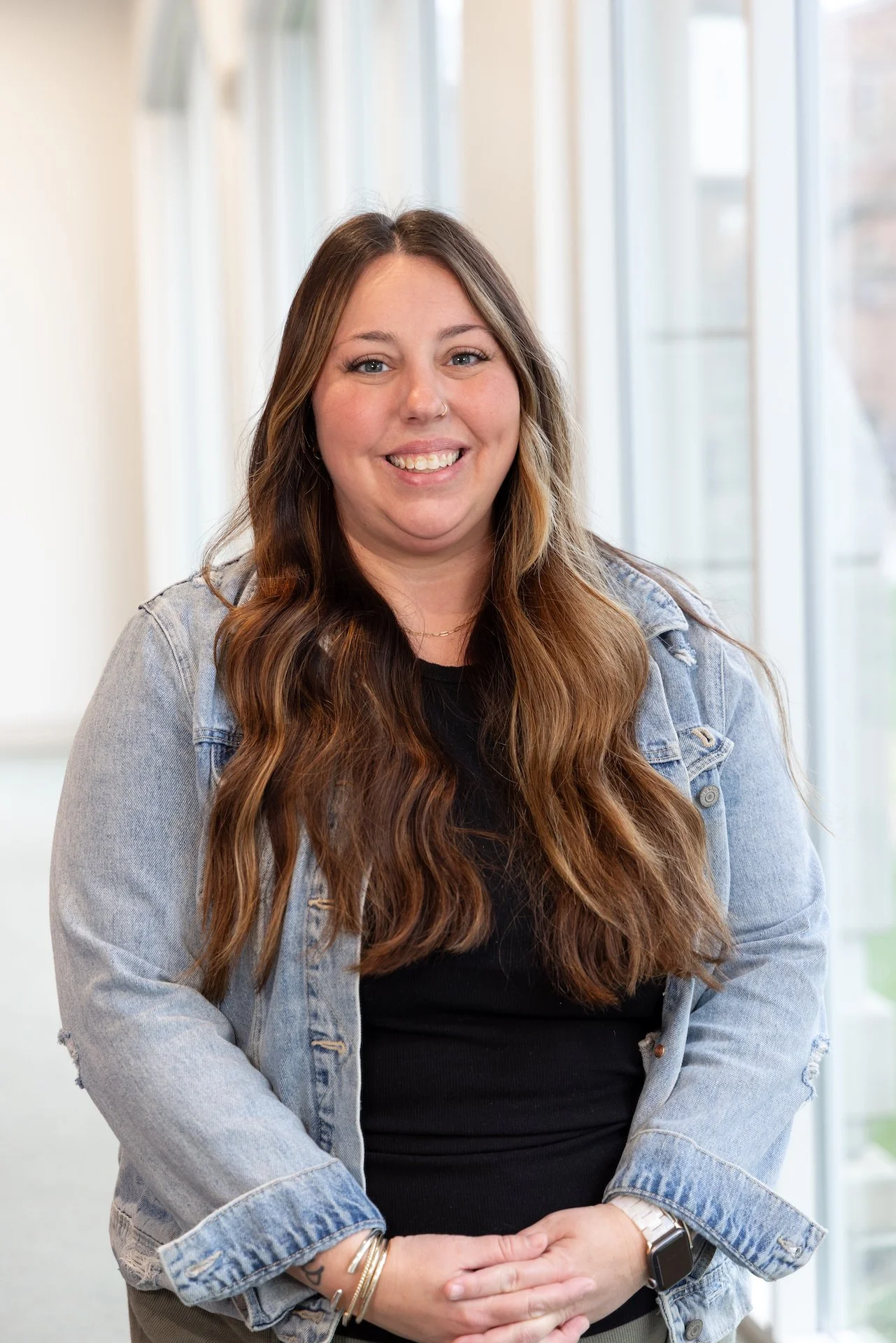 A woman with long wavy brown hair, wearing a denim jacket over a black top, smiling and standing indoors near large windows.