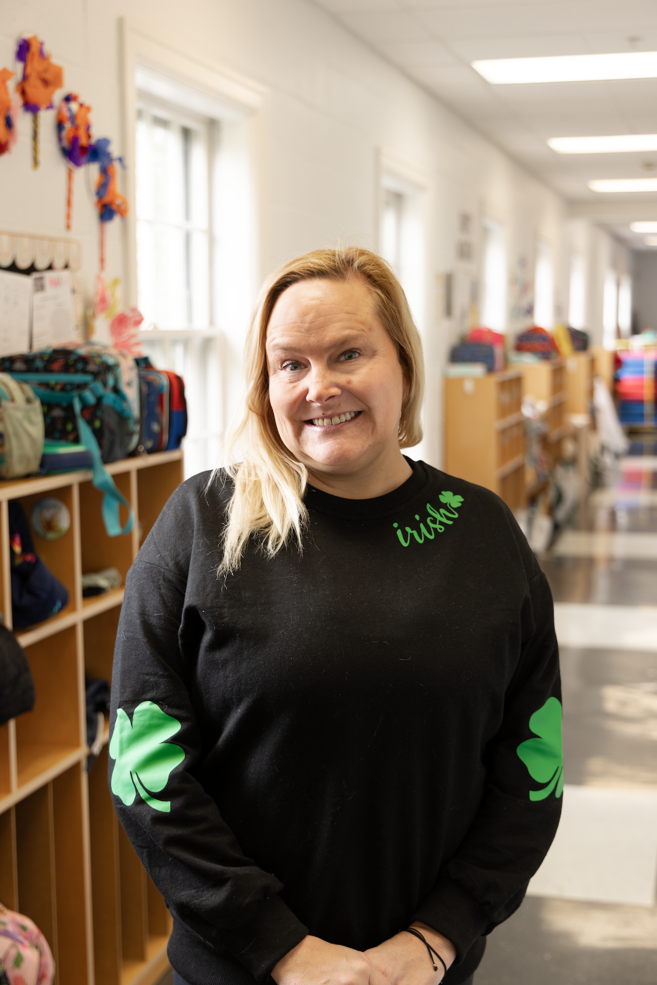 A woman with shoulder-length blonde hair smiling in a classroom, wearing a black sweatshirt with green shamrock designs and the word 'irish' on it.