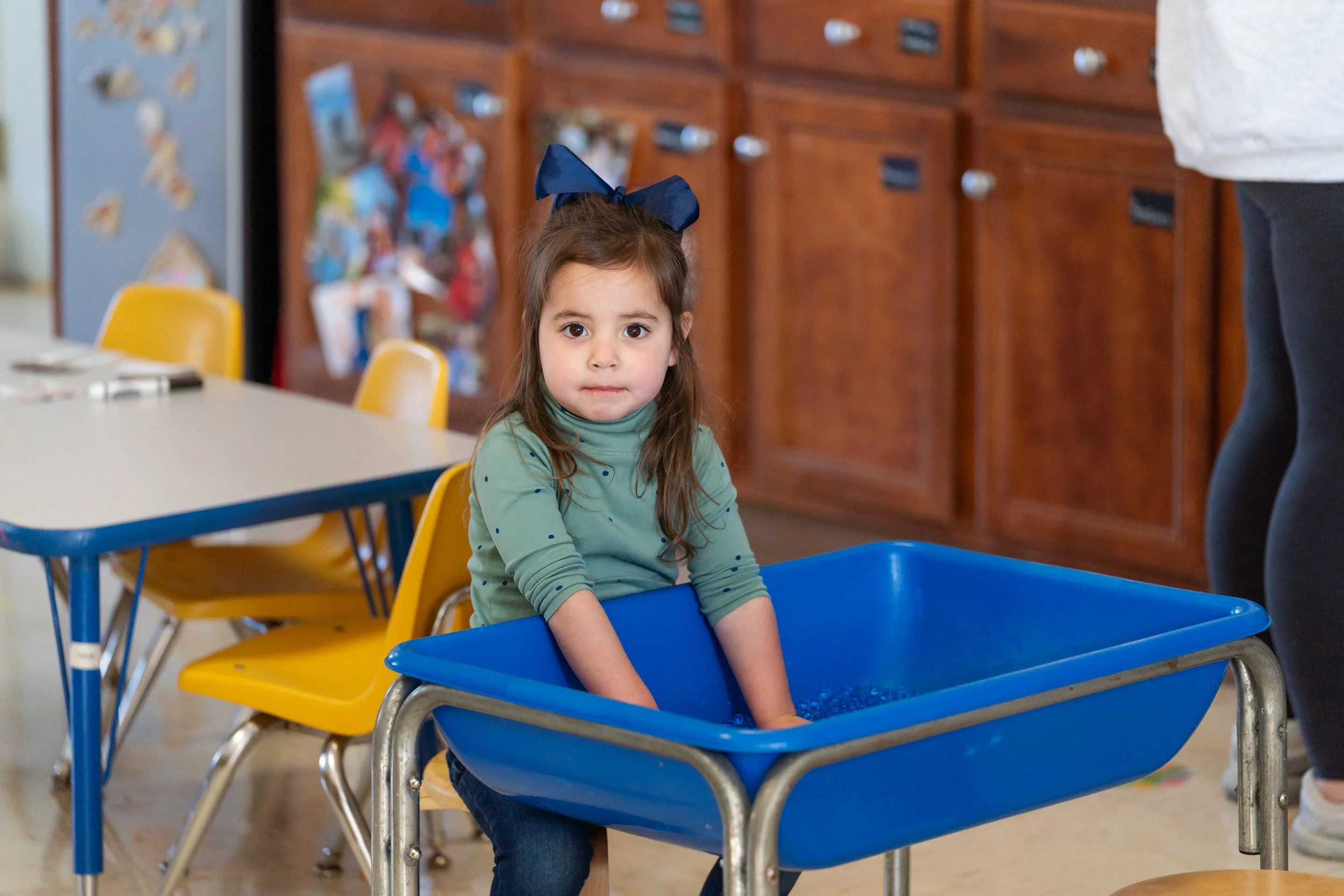 Young girl with long brown hair, wearing a green shirt with small dots and a large navy blue bow in her hair, standing in a blue plastic bin in a classroom.