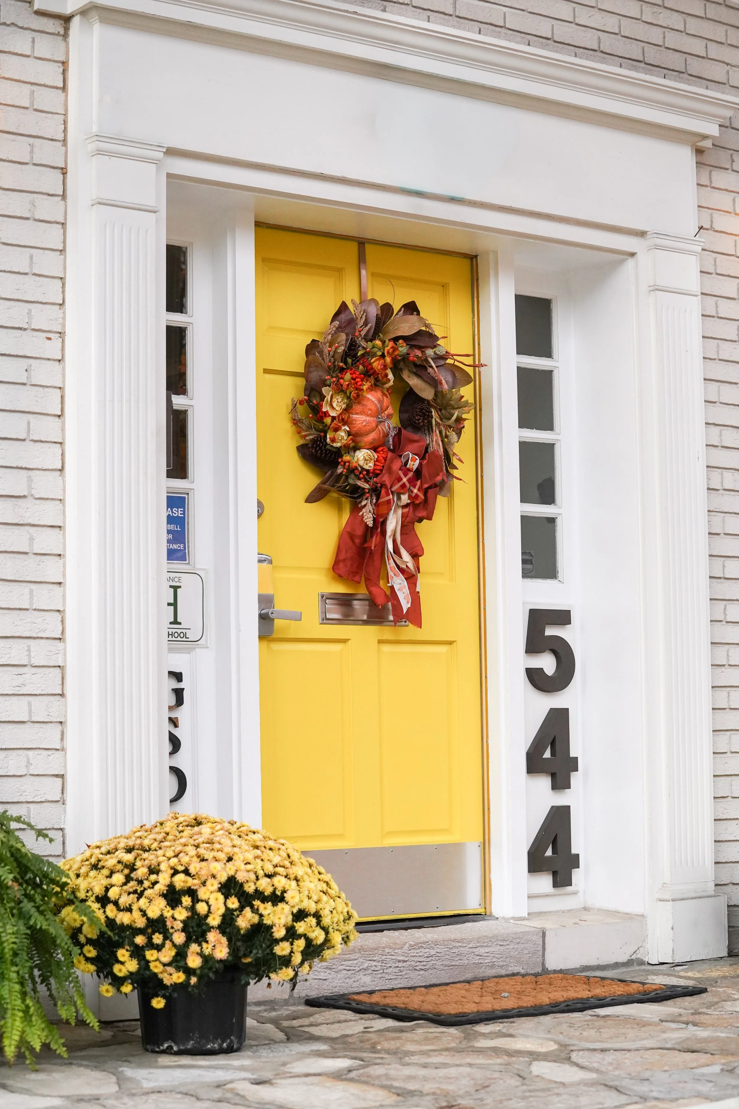 Yellow front door with fall wreath, house number 544, white brick exterior, potted yellow chrysanthemums, and a brown mat.