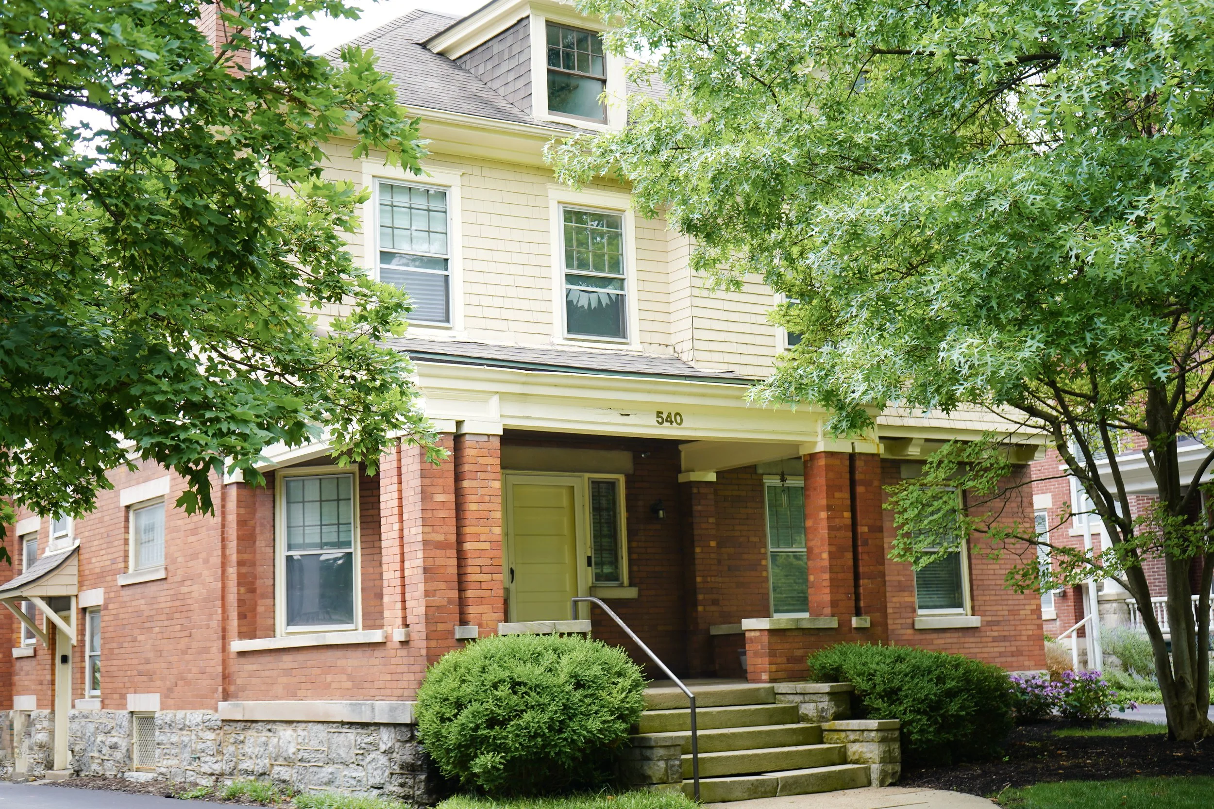 A residential house with a brick foundation, beige shingles, and a covered front porch, surrounded by green trees and bushes.