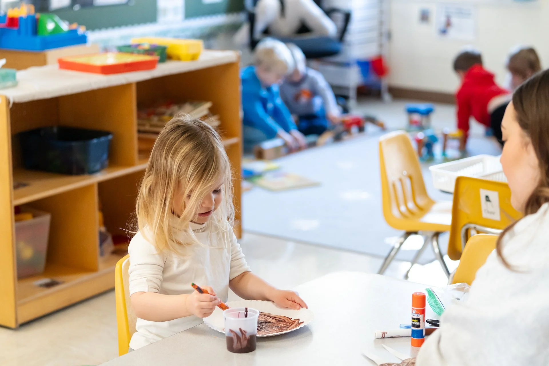 A young girl sits at a table in a classroom, painting on a paper plate with brown and green paint, with a brown cup of paint and glue nearby. In the background, other children are engaged in activities on the floor.