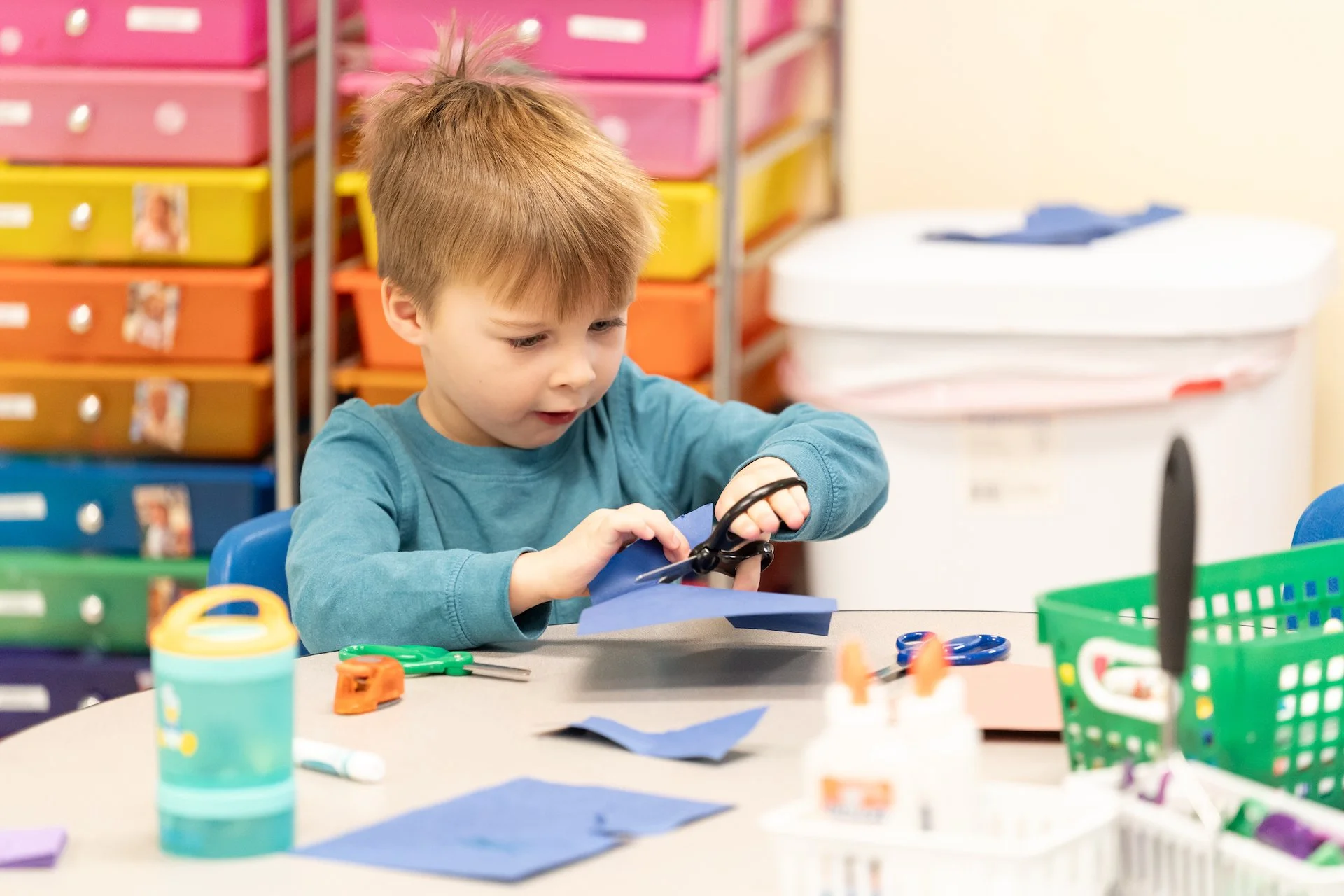 A young boy with red hair wearing a teal long-sleeve shirt, using scissors to cut blue paper at a table. The background shows colorful storage drawers and craft supplies.