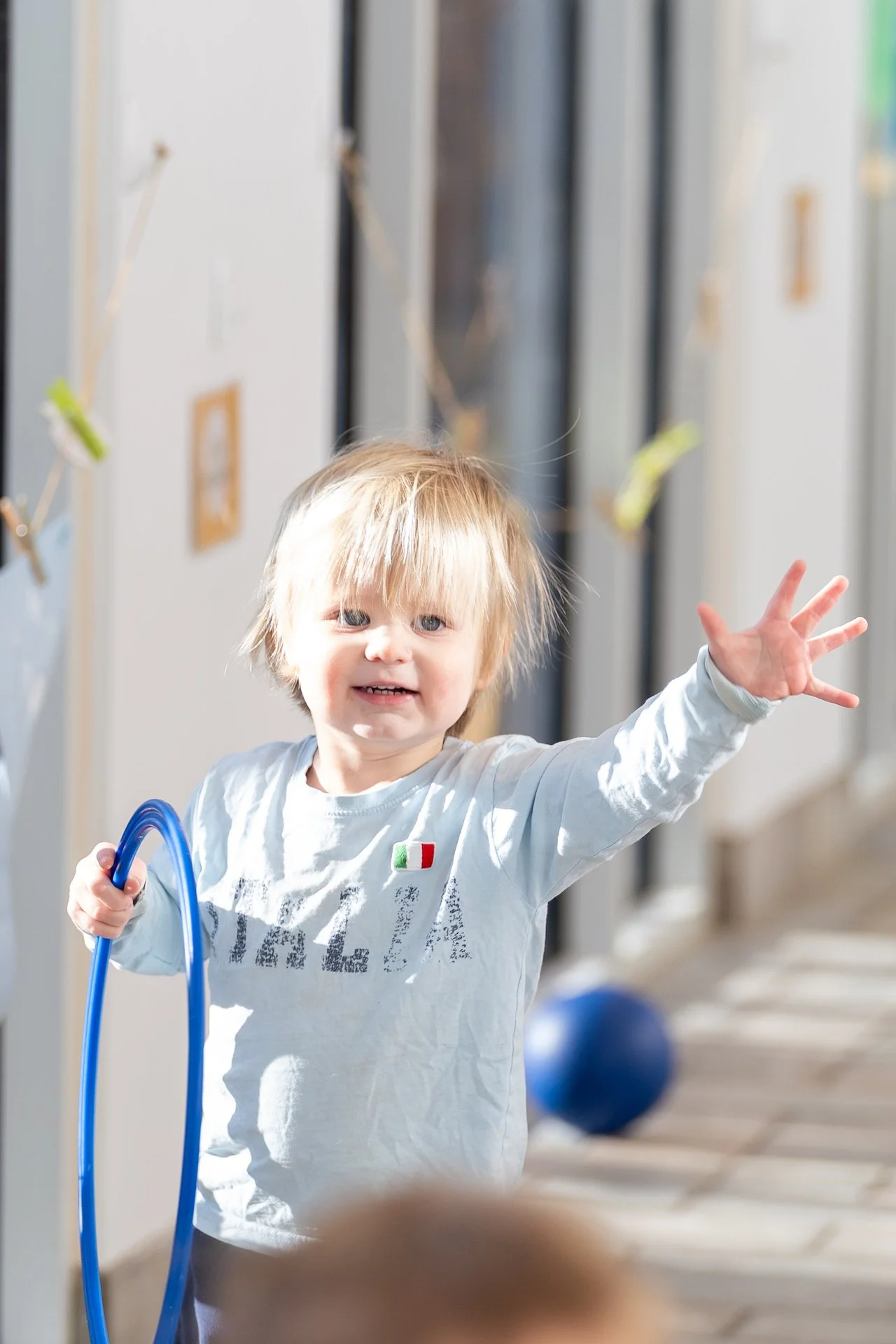 A young child with blonde hair and a white long-sleeve shirt reaching out with their left hand while holding a blue hula hoop in their right hand indoors, with lockers and a blue ball in the background.