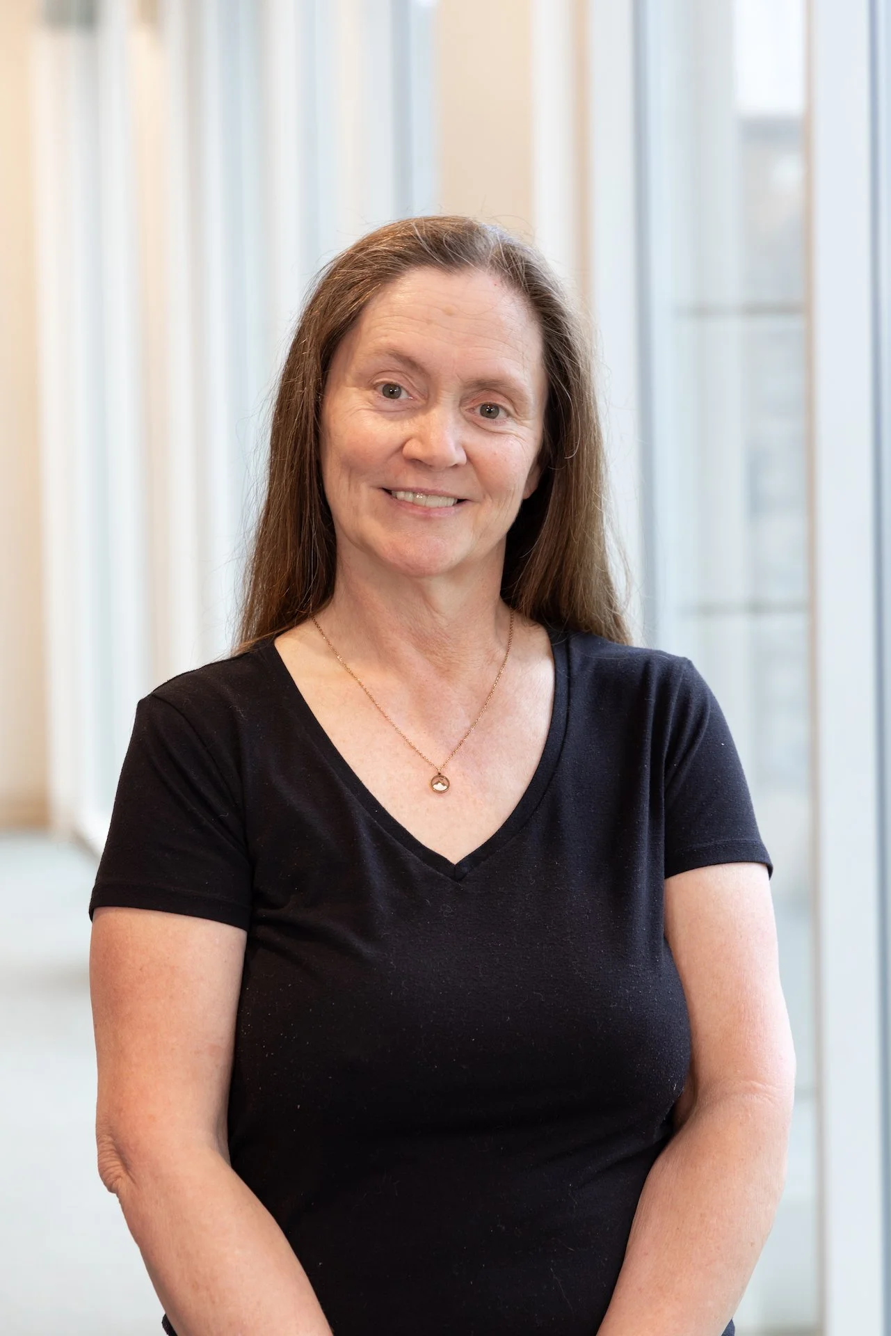 A middle-aged woman with long brown hair smiling, wearing a black V-neck T-shirt and a gold necklace, standing indoors near large windows.