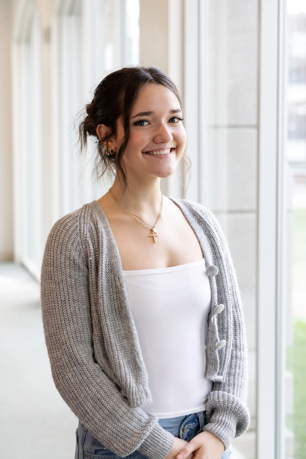 A young woman standing indoors near large windows, smiling at the camera. She has dark hair tied back, wears earrings, a gold necklace with a cross pendant, a white tank top, and a gray knitted cardigan.