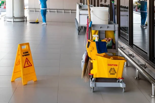 Janitorial cleaning supplies and equipment, including a yellow caution sign, mop bucket, and cleaning tools, in an airport corridor.
