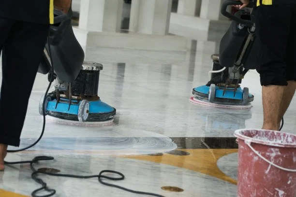 Workers cleaning or polishing a large floor using floor machines with blue pads, with buckets nearby.