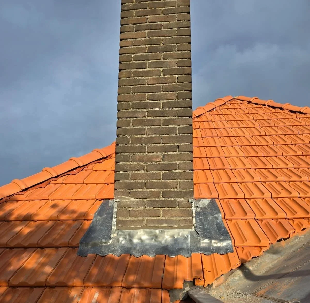 Tegelbedekte dakrand met een stenen schoorsteen en een hemel met bewolking.