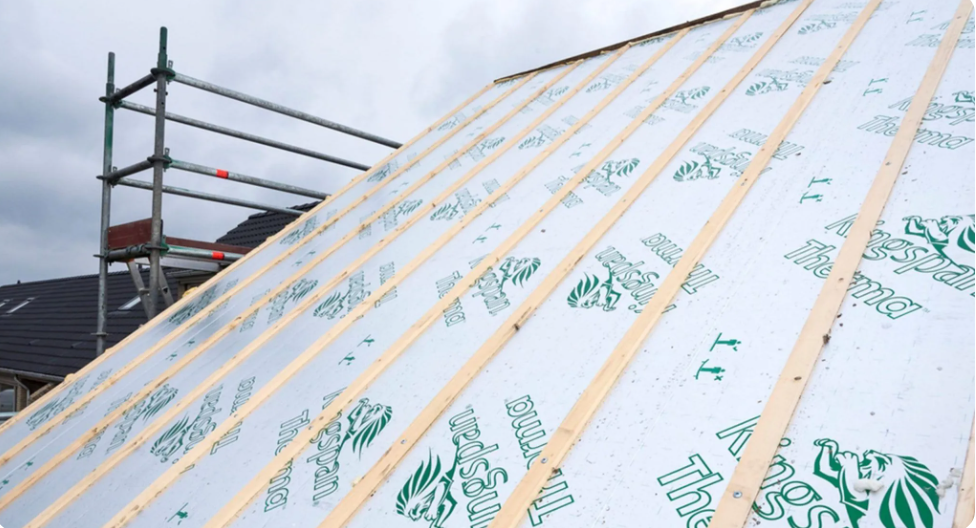 Dakbedekking in aanbouw met houten latten en waterdichte wrap op een schuin dak, met een bouwsteiger op de achtergrond onder een bewolkte hemel.