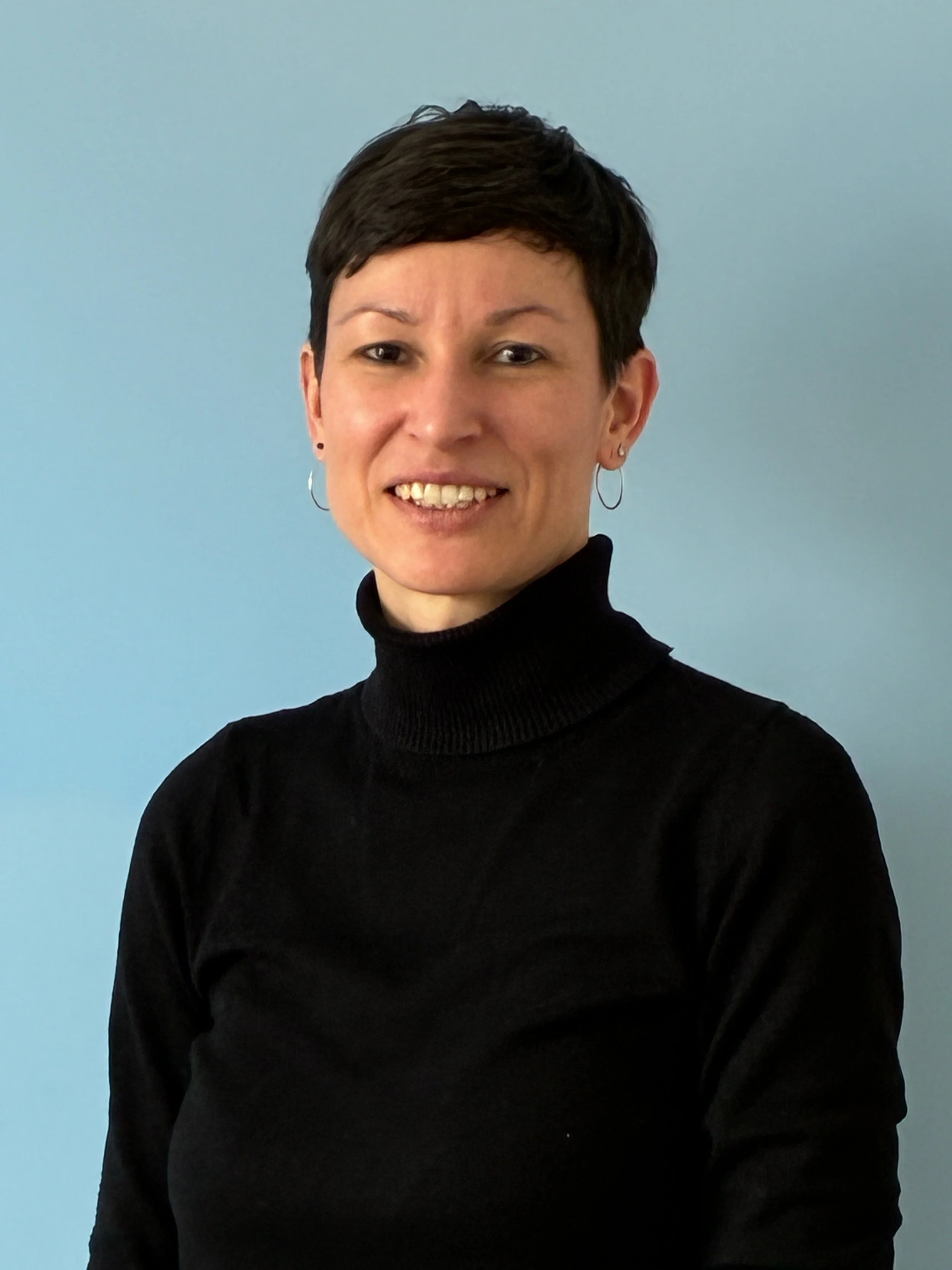 Eine Frau mit Kurzhaarfrisur, schwarzen Turtleneck und Ohrringen, lächelnd vor pastellblauem Hintergrund.