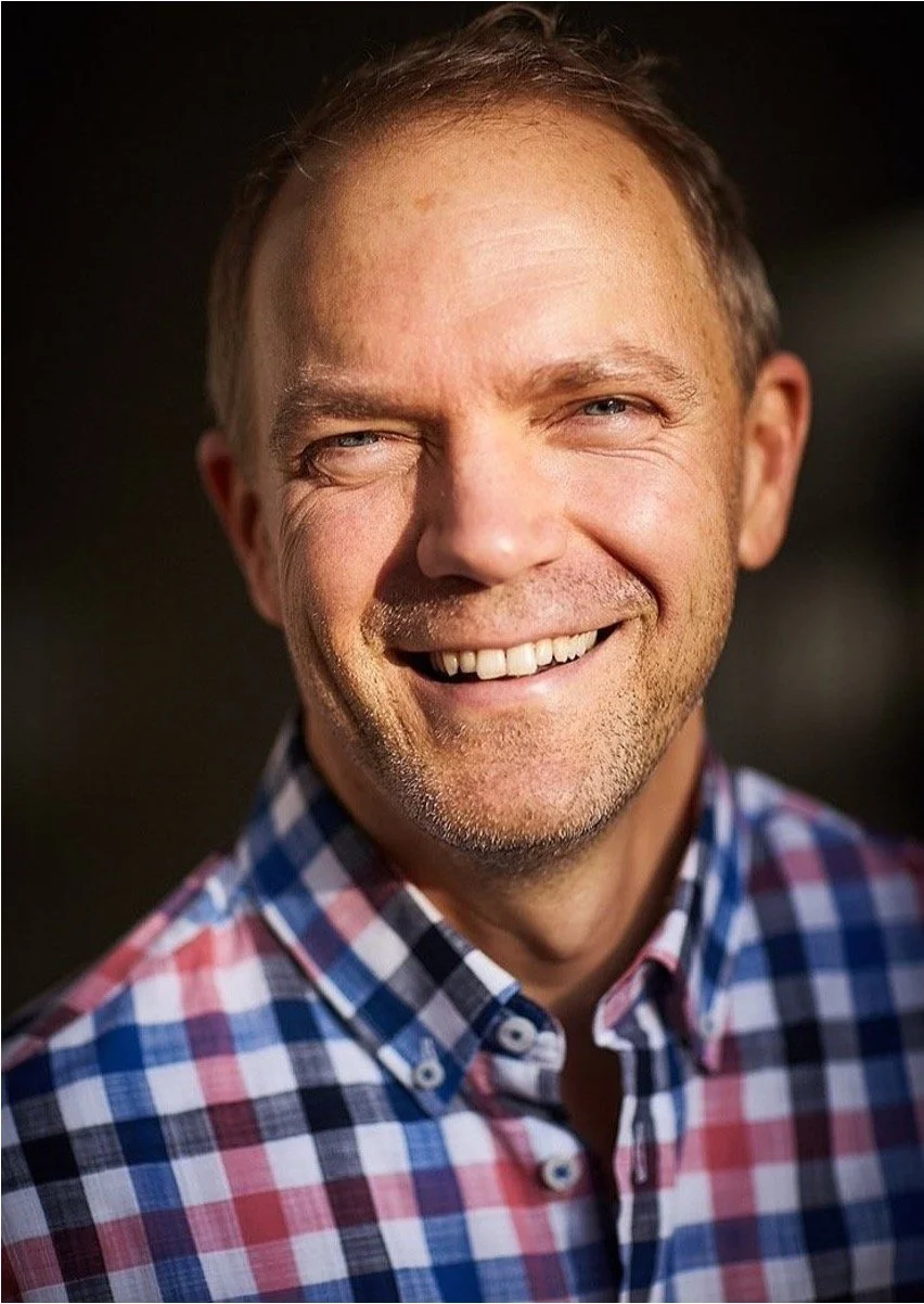 Ein lachender Mann mit kurzen Haaren, Bart und einem karierten Hemd, im Freien bei Sonnenlicht fotografiert.