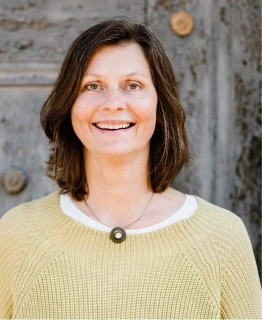 Eine Frau mit braunen, schulterlangen Haaren, die lächelnd nach vorne schaut, vor einem holzverkleideten Hintergrund.