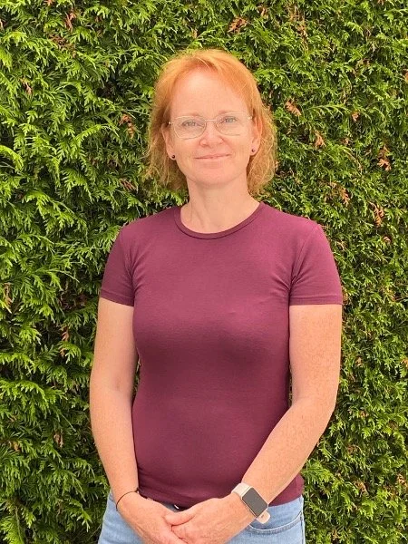 Eine Frau mit roten Haaren, Brille und einem lila T-Shirt steht vor einer grünen Hecke.