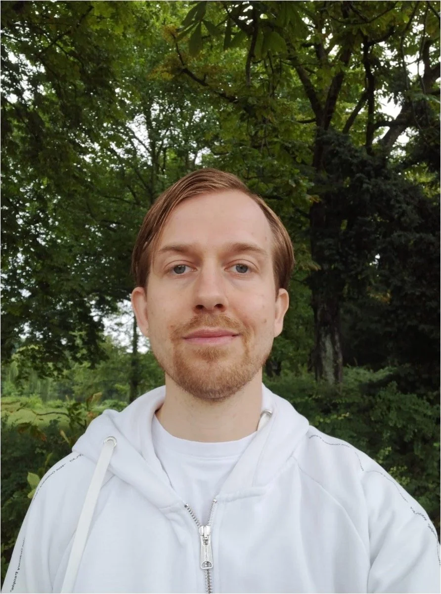 Nahaufnahme eines Mannes mit braunem Haar und Bart, der vor einem grünen, bewaldeten Hintergrund steht, trägt ein weißes Sweatshirt.