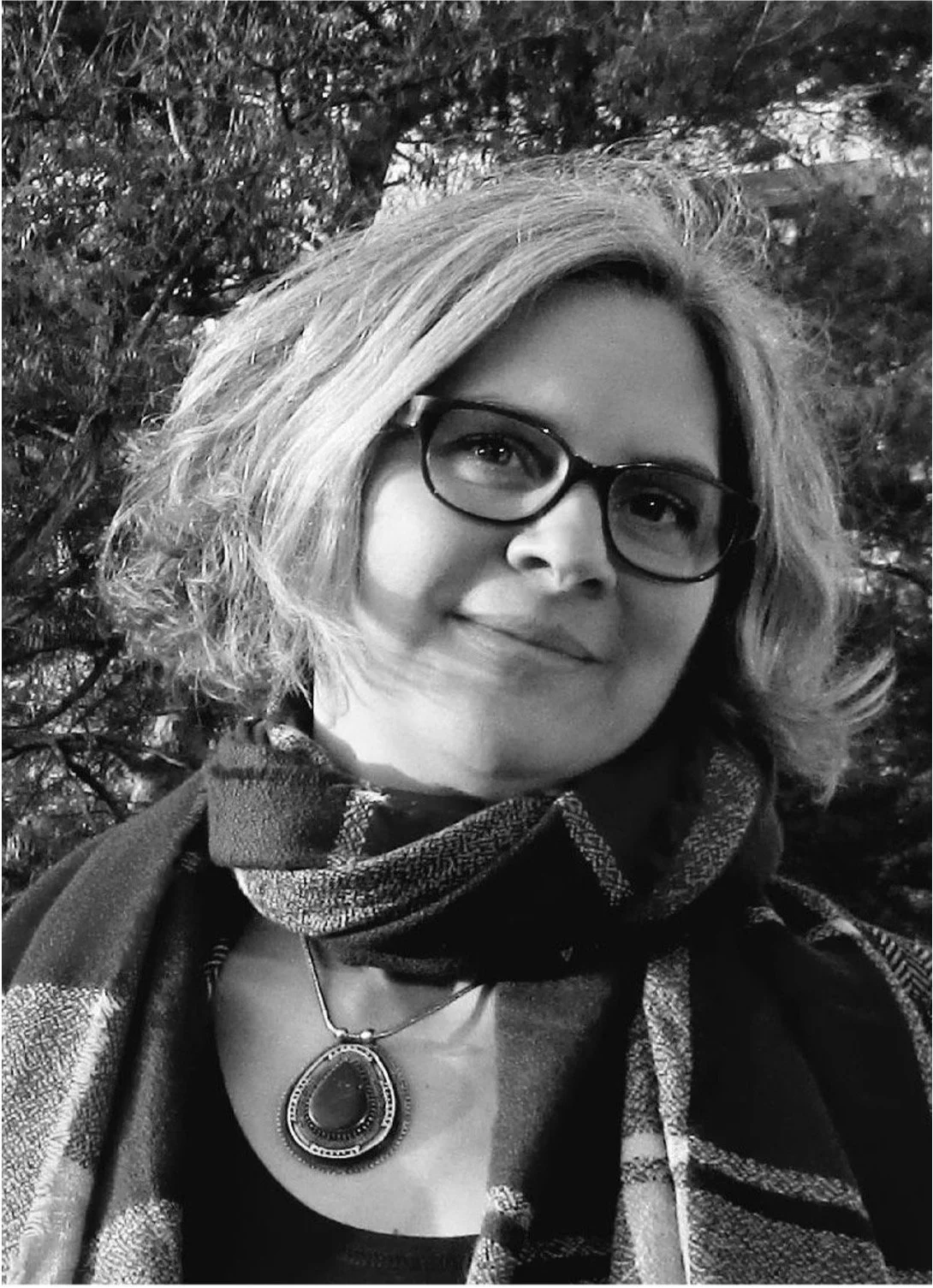 DamenPortrait, kurz gewelltes Haar, Brille, Halskette, Schal, lächelnd, outdoor, Bäume im Hintergrund, Schwarz-Weiß-Foto.