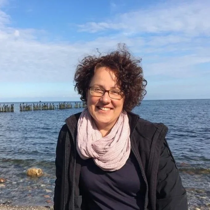 Eine lachende Frau mit lockigem Haar steht am Strand vor dem Meer, im Hintergrund sind Steine und ein Himmel mit wenigen Wolken.