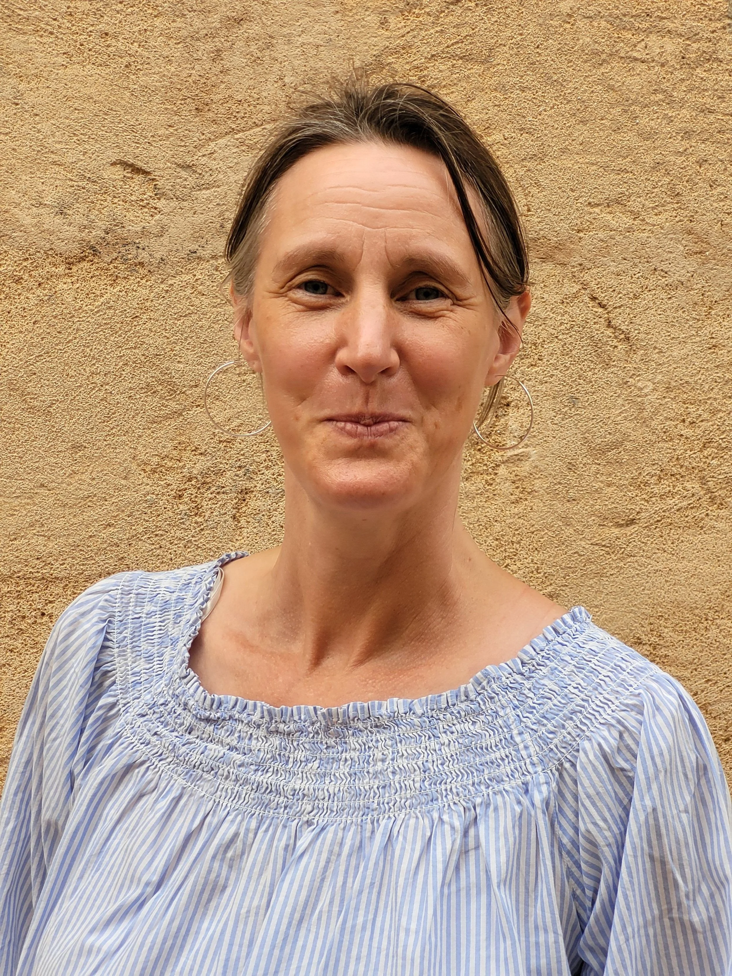 Frau mit kurzen braunen Haaren, kreisförmigen Ohrringen, im blauen Kleid vor einer terrakottafarbenen Wand.