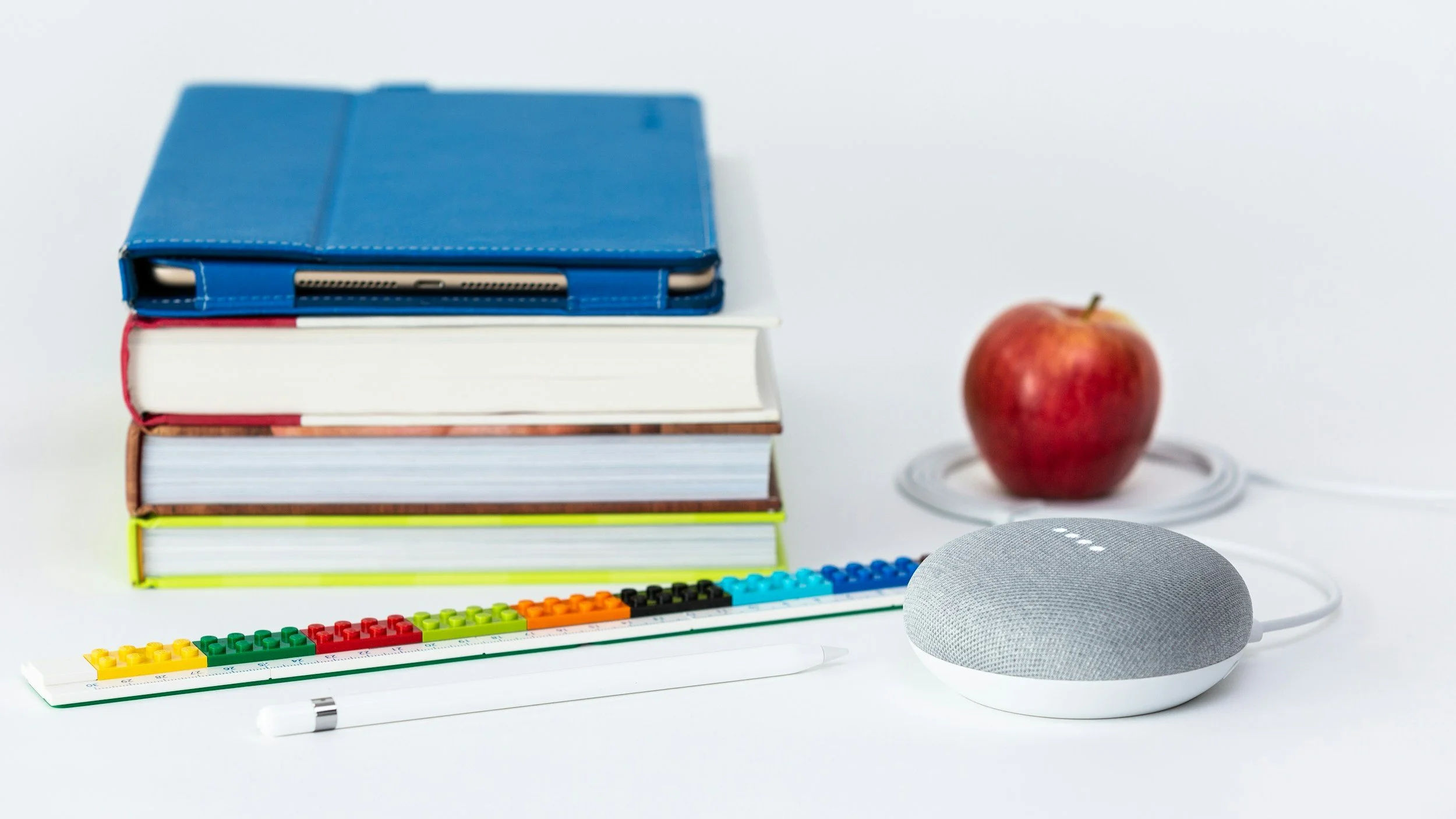 Stack of books with a blue tablet case on top, an apple, a Google Nest speaker, a rainbow-colored list of domino-like pegs, and a red pen on a white surface.