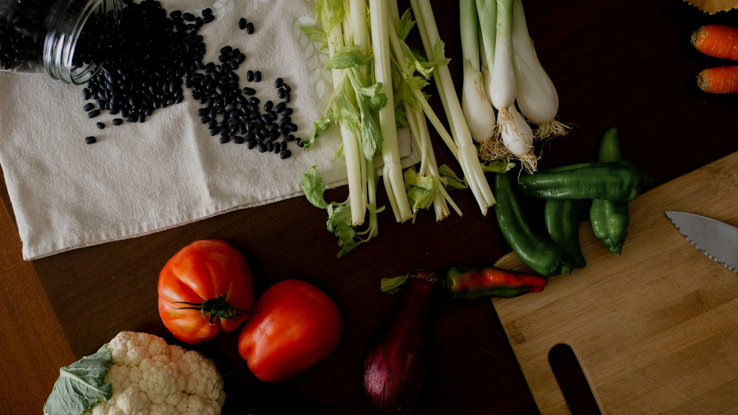 Fresh vegetables including tomatoes, cauliflower, black beans, celery, white onions, jalapeño peppers, carrots, and red onion on a wooden table with a cutting board and knife.