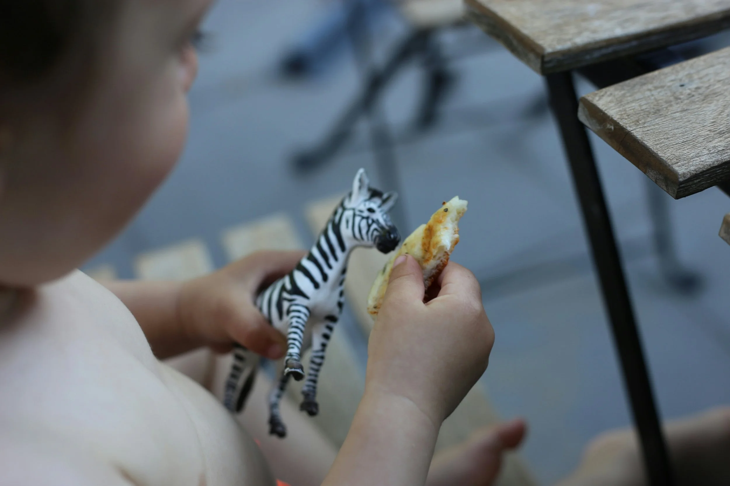 A young child holding a small zebra figurine in one hand and a slice of pizza in the other, sitting next to a wooden table.