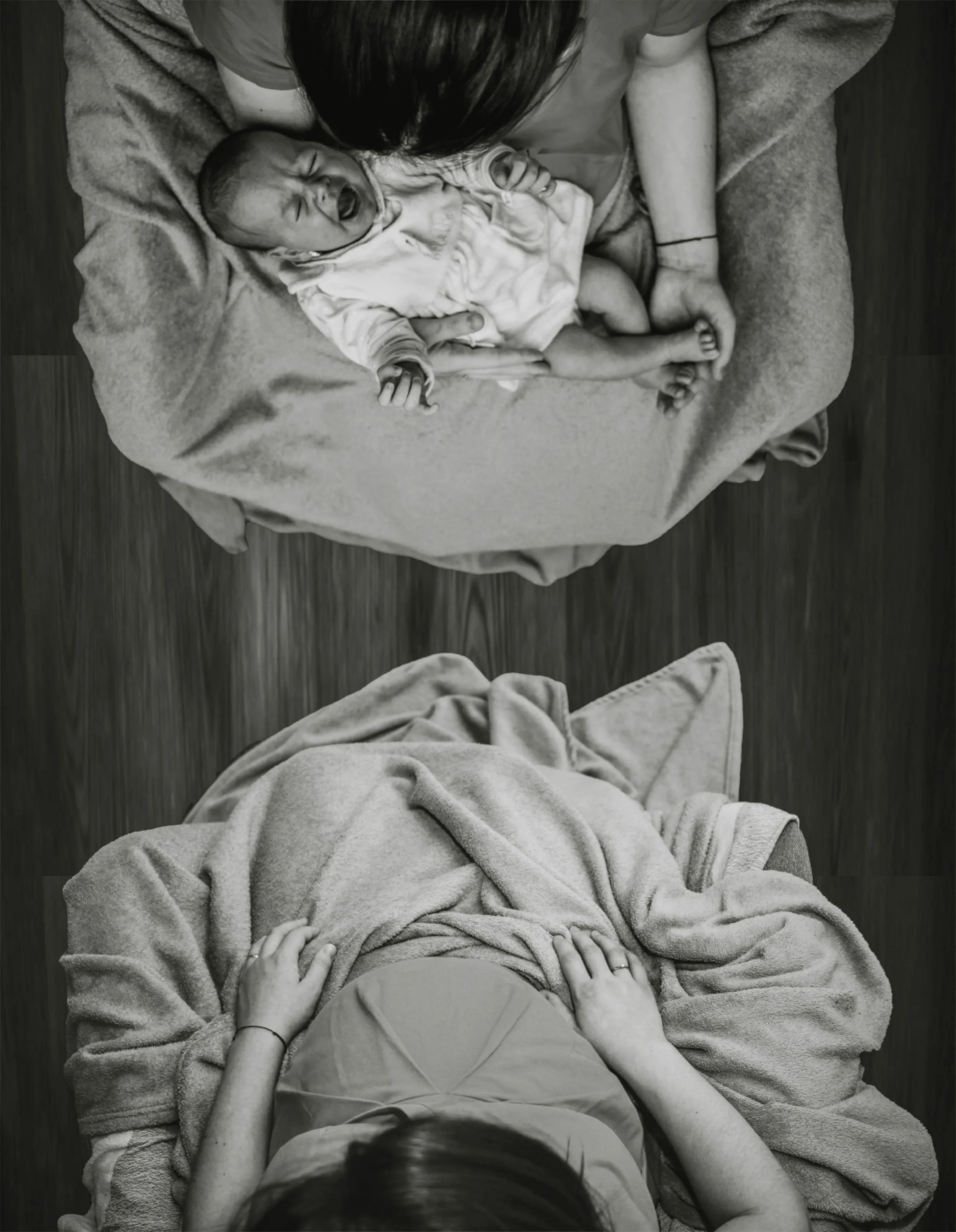 Top view of a person lying in bed with their hands on their stomach, covered with a blanket, looking down, and another person holding a crying baby in their arms, with the baby's head resting on their arm.