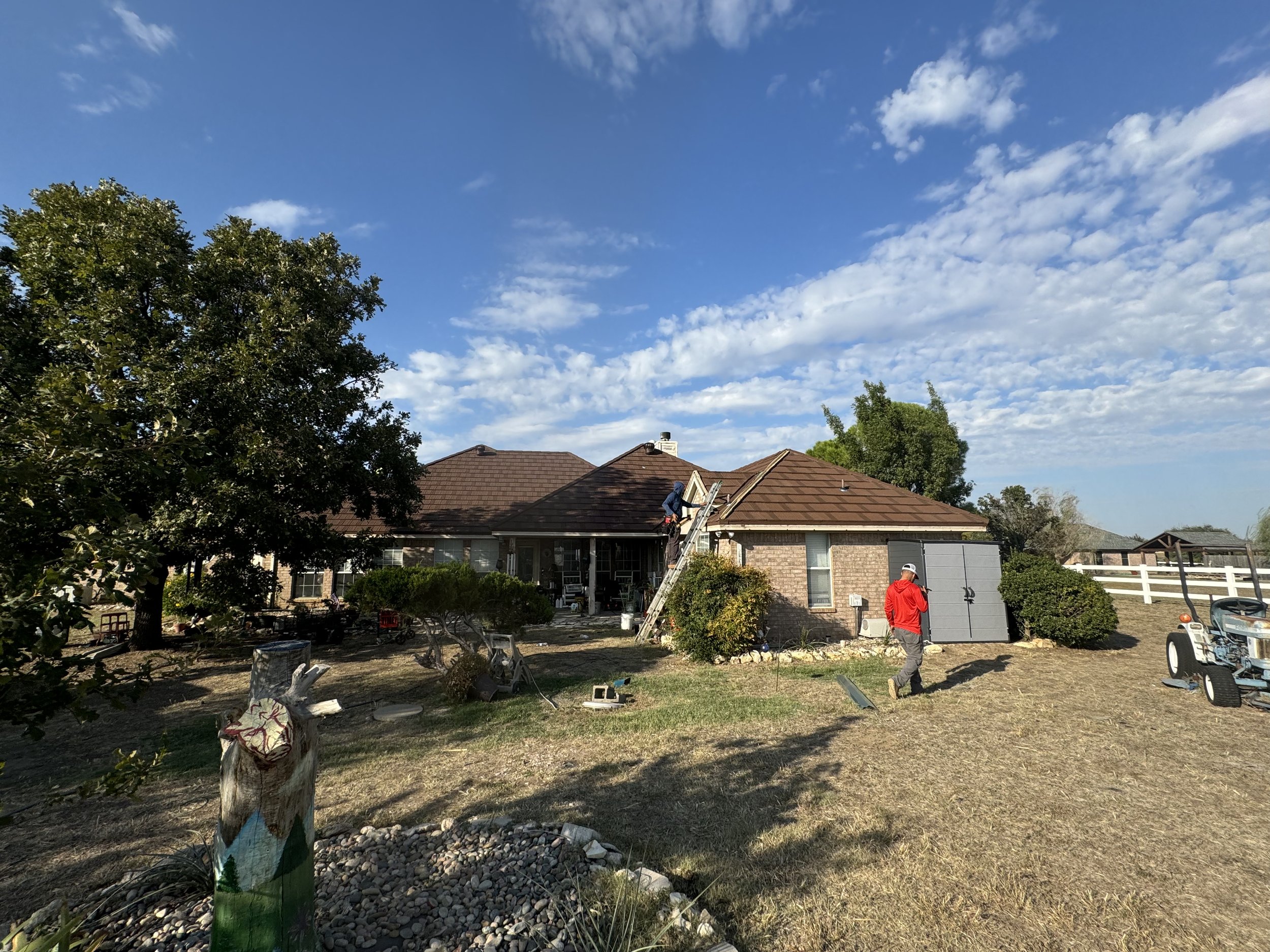 Residential backyard with workers installing a roof on a single-story brick house under a partly cloudy sky.