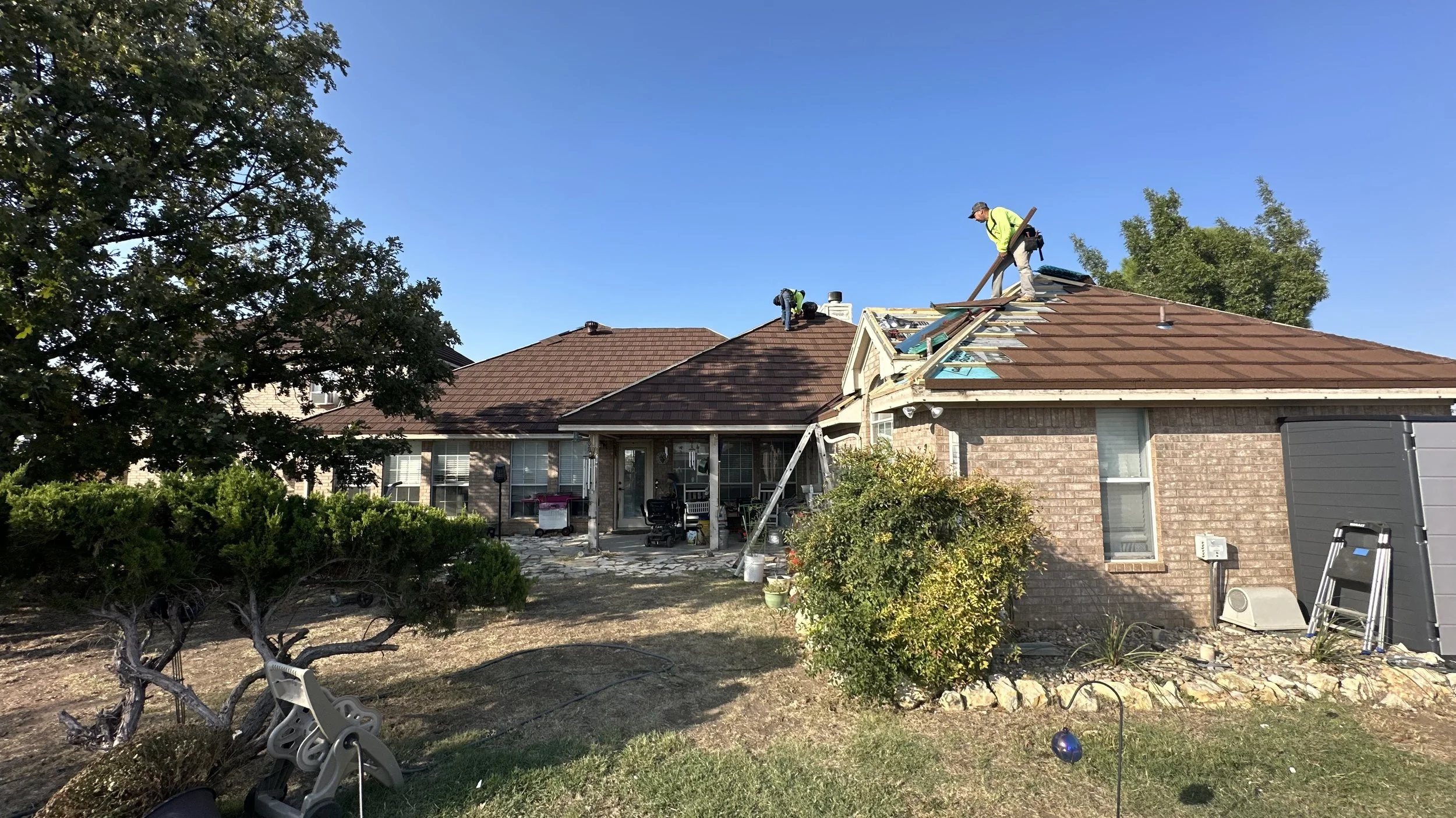 Three construction workers repairing or replacing the roof of a brick house. One worker is on the roof holding a wooden beam, another climbing with a ladder, and the third is on the roof working near the chimney. The backyard has a tree, bushes, and 