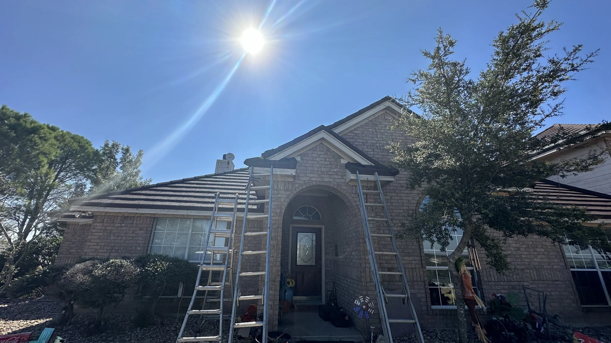 Front view of a brick house with two ladders leaning against the entrance, a tree on the right, and the sun shining brightly in a clear blue sky.