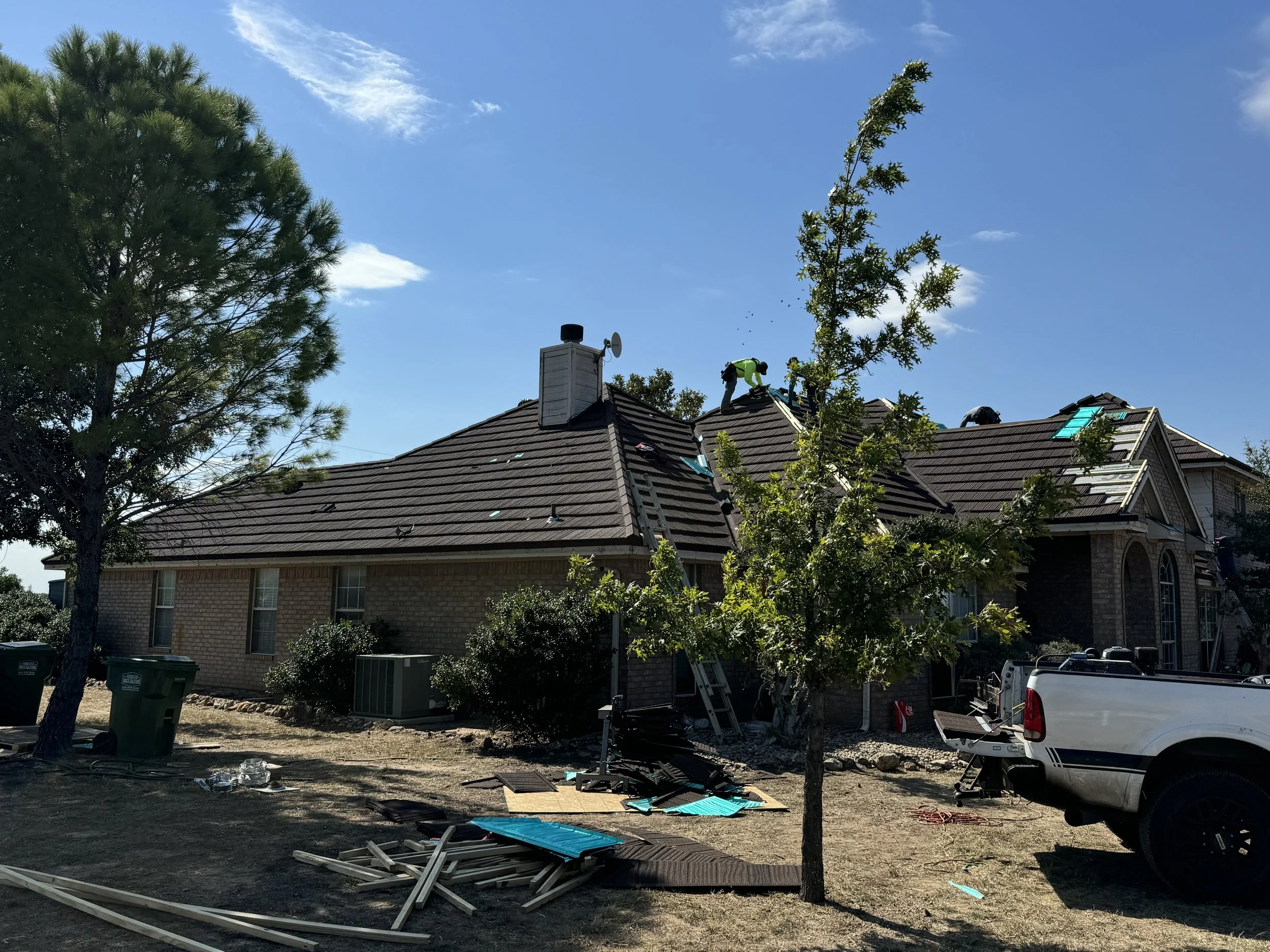 Construction workers are repairing or installing roofing on a large brick house during the day. Debris and tools are scattered on the ground, and a construction truck is parked nearby.