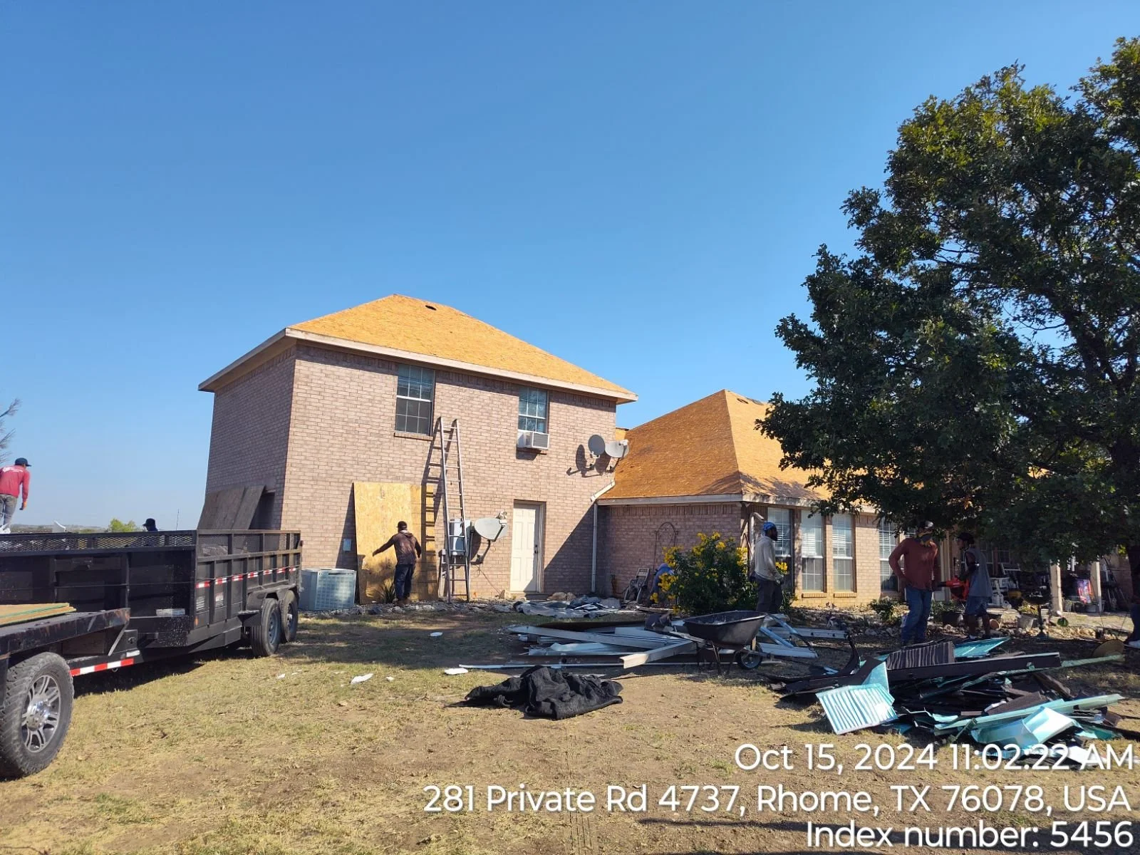 Workers demolishing a brick house with yellow roofing shingles on a sunny day, debris scattered on the ground, a trailer parked nearby, and a large tree in the yard.