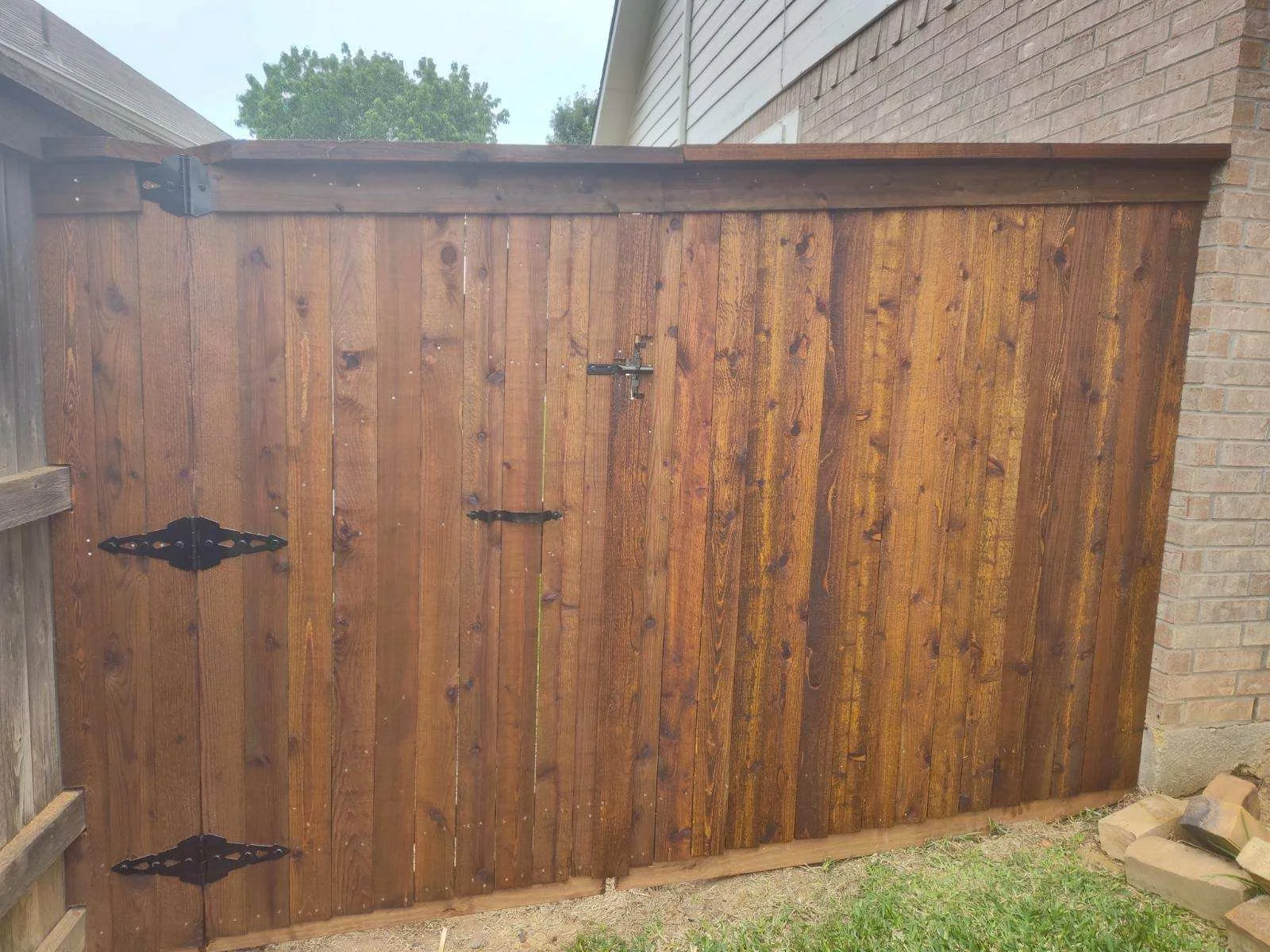Newly installed wooden backyard gate with decorative black metal hinges and latch, adjacent to brick house and second fence post, with grass and bricks at the base.
