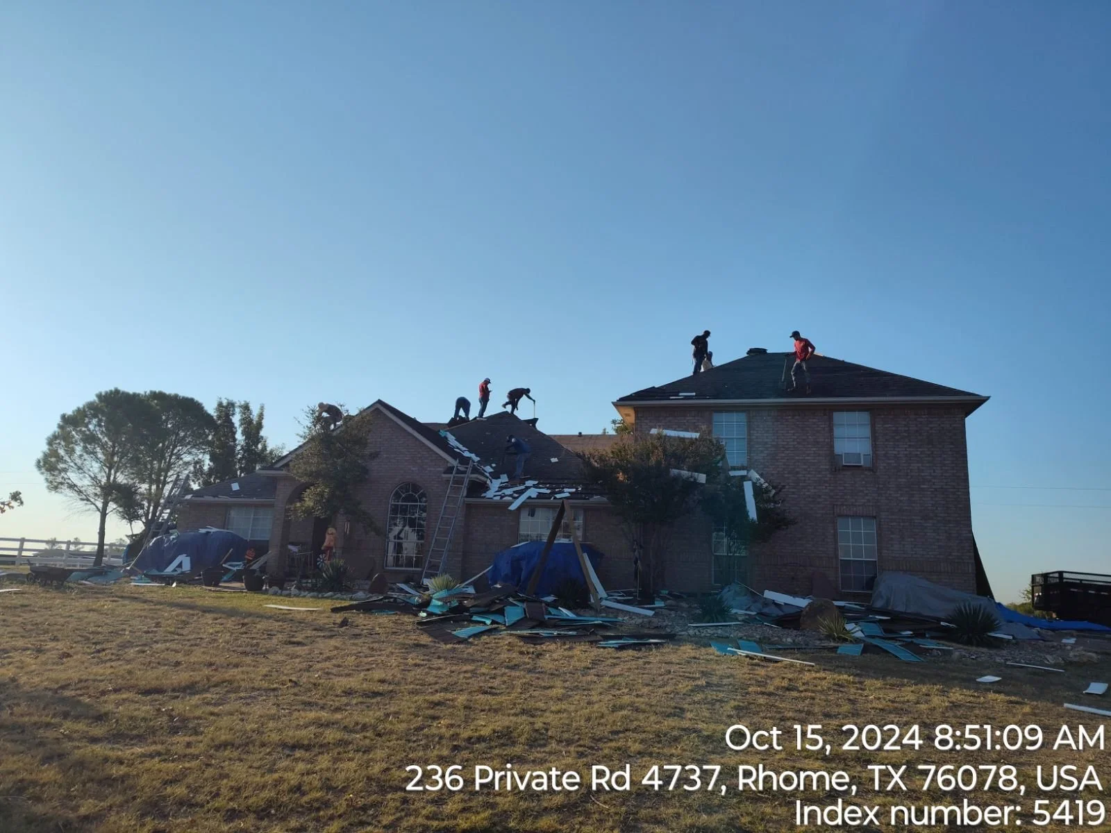 Workers on the roof of a brick house repairing damage, debris scattered at the yard, some covered with blue tarps, with the time and location details overlaid.