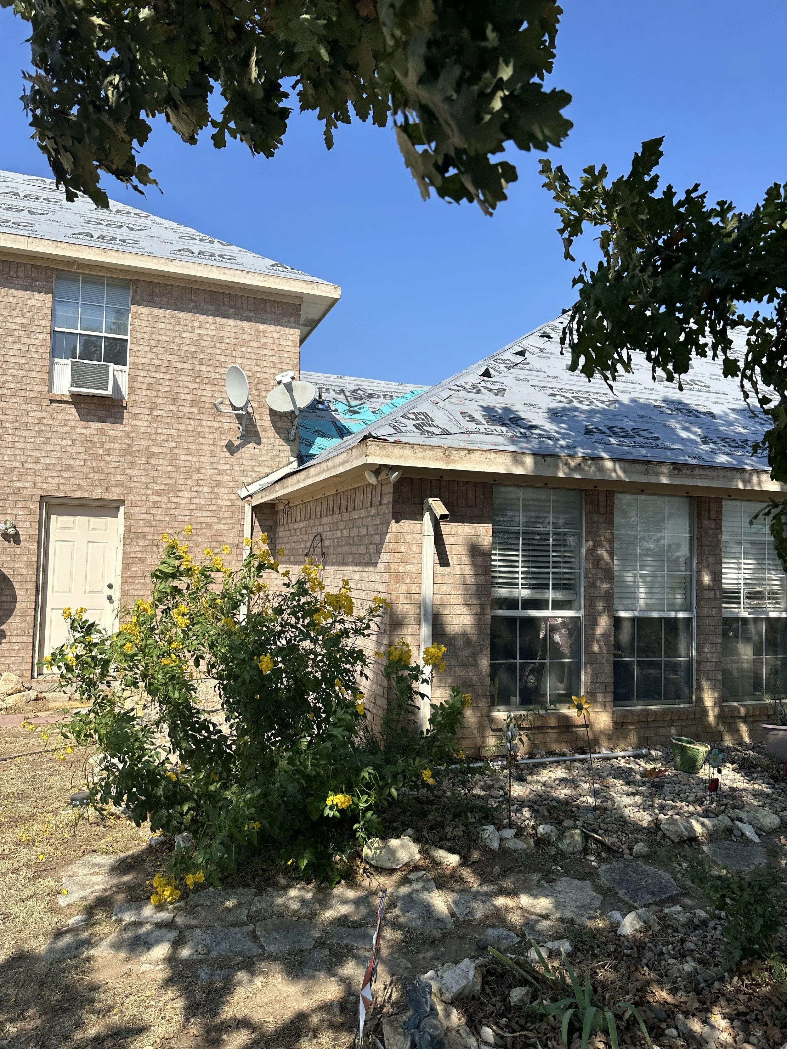 A house under construction or renovation with a shingled roof and brick exterior. The roof appears to be partially covered with weatherproofing material. There are two satellite dishes on the side of the house, and a small garden with yellow flowers 