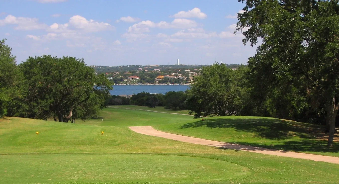 A golf course with a green fairway, trees on both sides, a sand trap, and a walking path. In the background, there is a body of water and a city skyline with buildings under a partly cloudy sky.