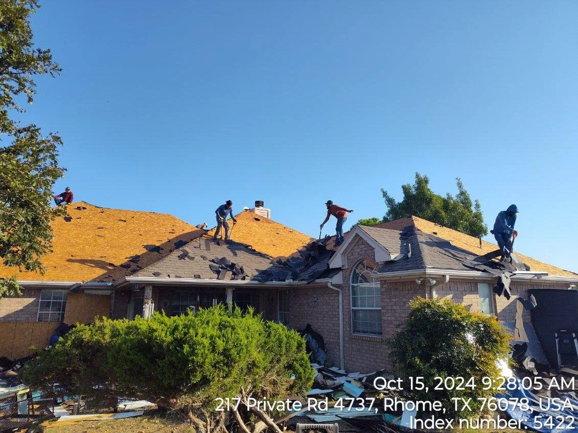 Four workers are on the roof of a house dismantling and removing shingles during the daytime. Debris and shingles are scattered across the roof. The house has brick walls and a front porch. The scene takes place in Rhome, Texas, on October 15, 2024.