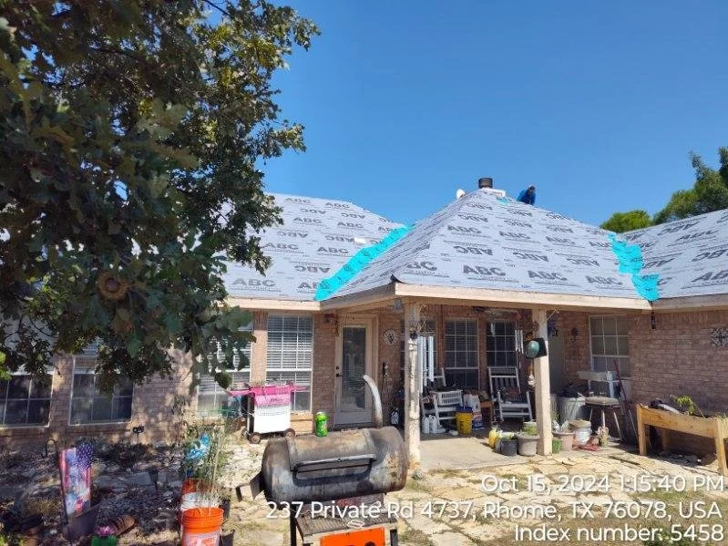 A house under construction with roofing materials, a porch with chairs, and various tools and containers in the yard, under a clear blue sky.