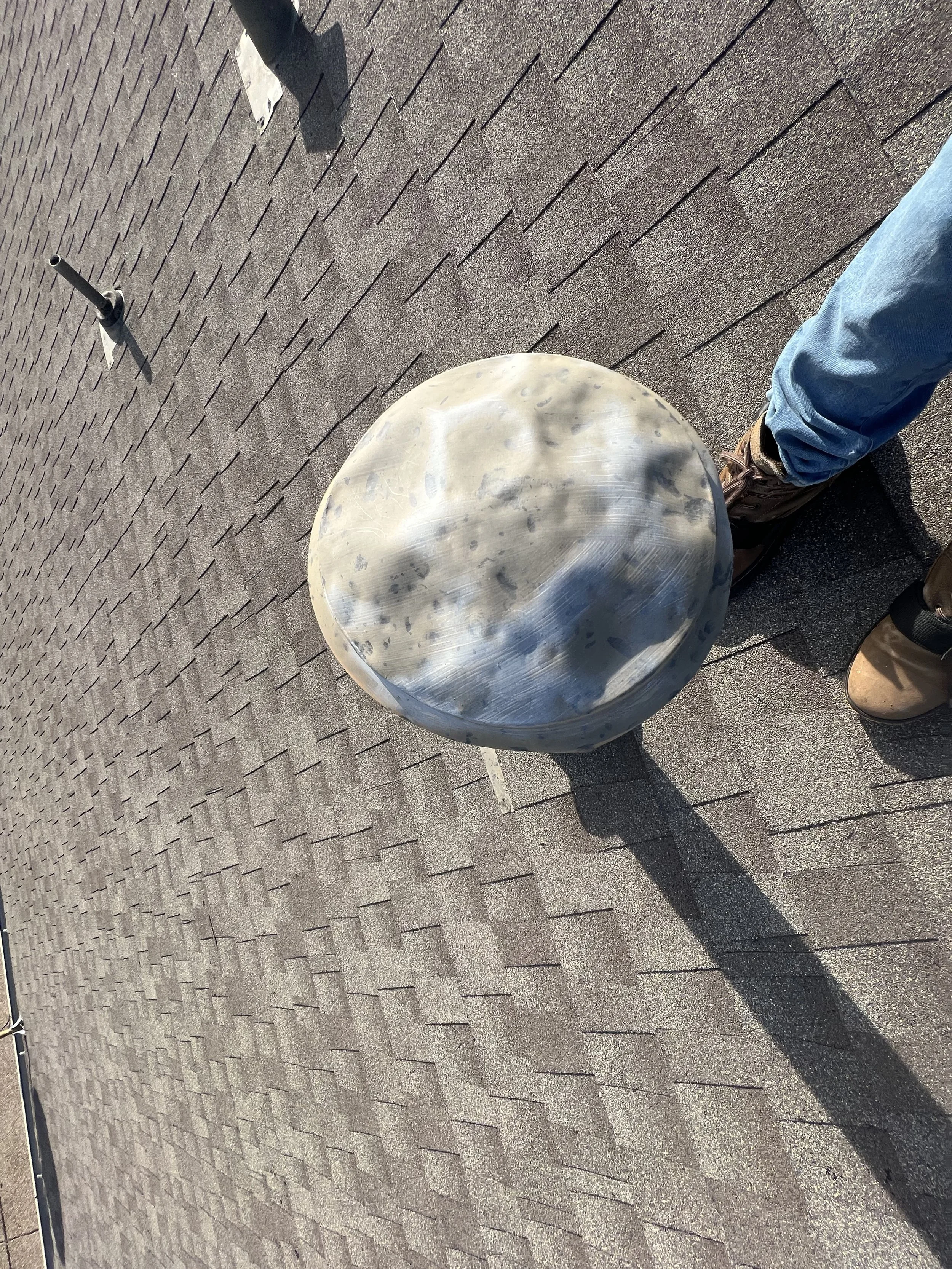 Close-up view of a metal vent pipe on a shingled roof, with a person’s foot and part of their leg visible nearby.